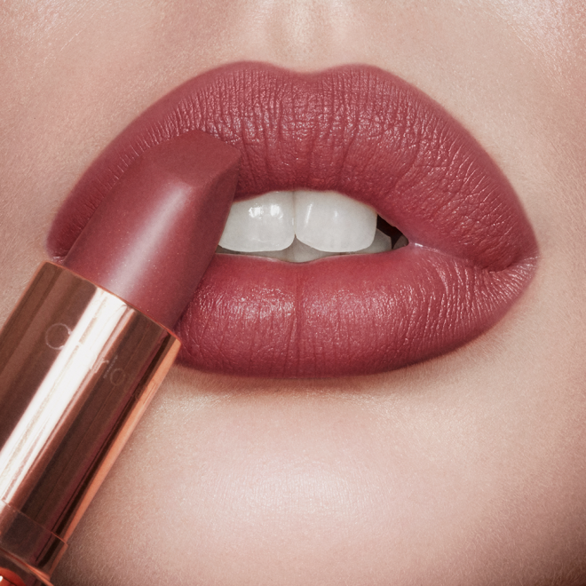 Lips close-up of a light-tone model wearing a warm, berry-pink matte lipstick while holding the lipstick in front of her lips. 