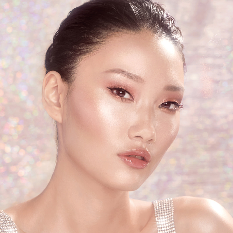 A fair-tone brunette model wearing soft pink eye makeup with nude pink lip gloss and glowy pink blush with an extremely glowy and flawless face base. 