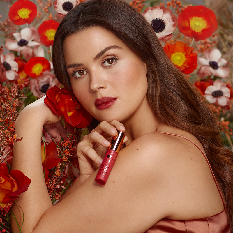 A medium-tone brunette model with brown eyes wearing a soft, peachy-brown lip tint and holding the lip tint in one hand and a flower in the other.