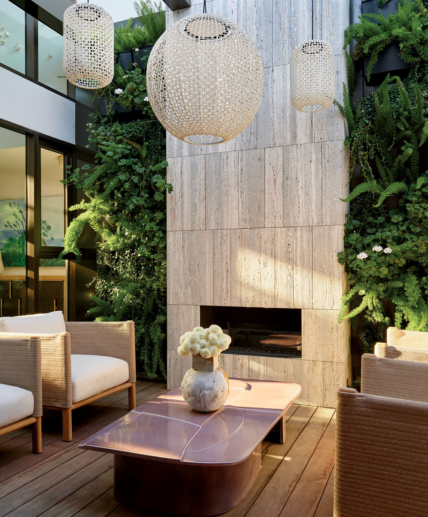 Stylish patio with woven chairs, a pink coffee table, and a tall stone fireplace. Lush greenery surrounds, accented by elegant hanging lanterns.