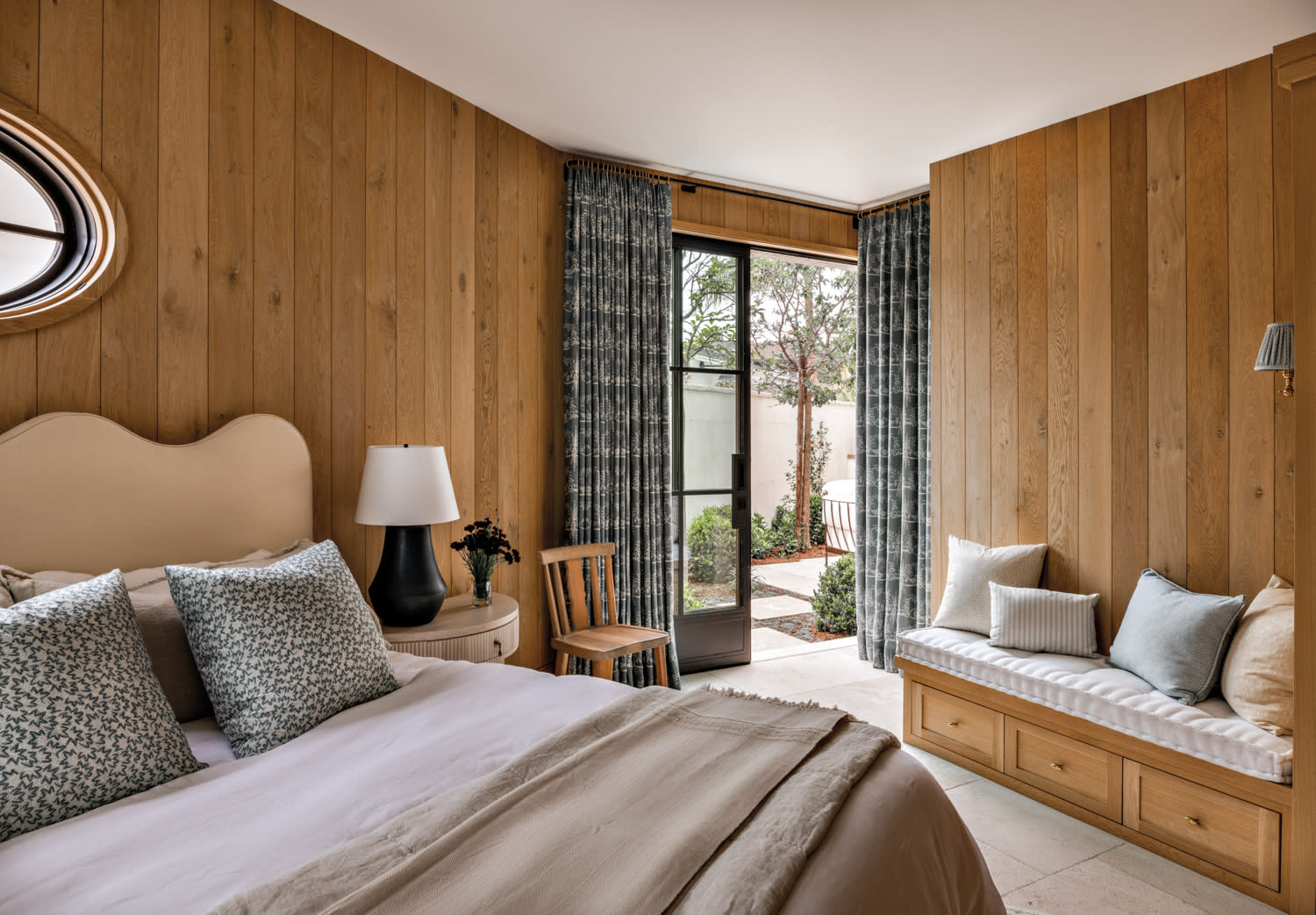 bedroom with oak paneled walls, bed with wavy upholstered headboard, and black and glass steel doors in home by Marie Carson