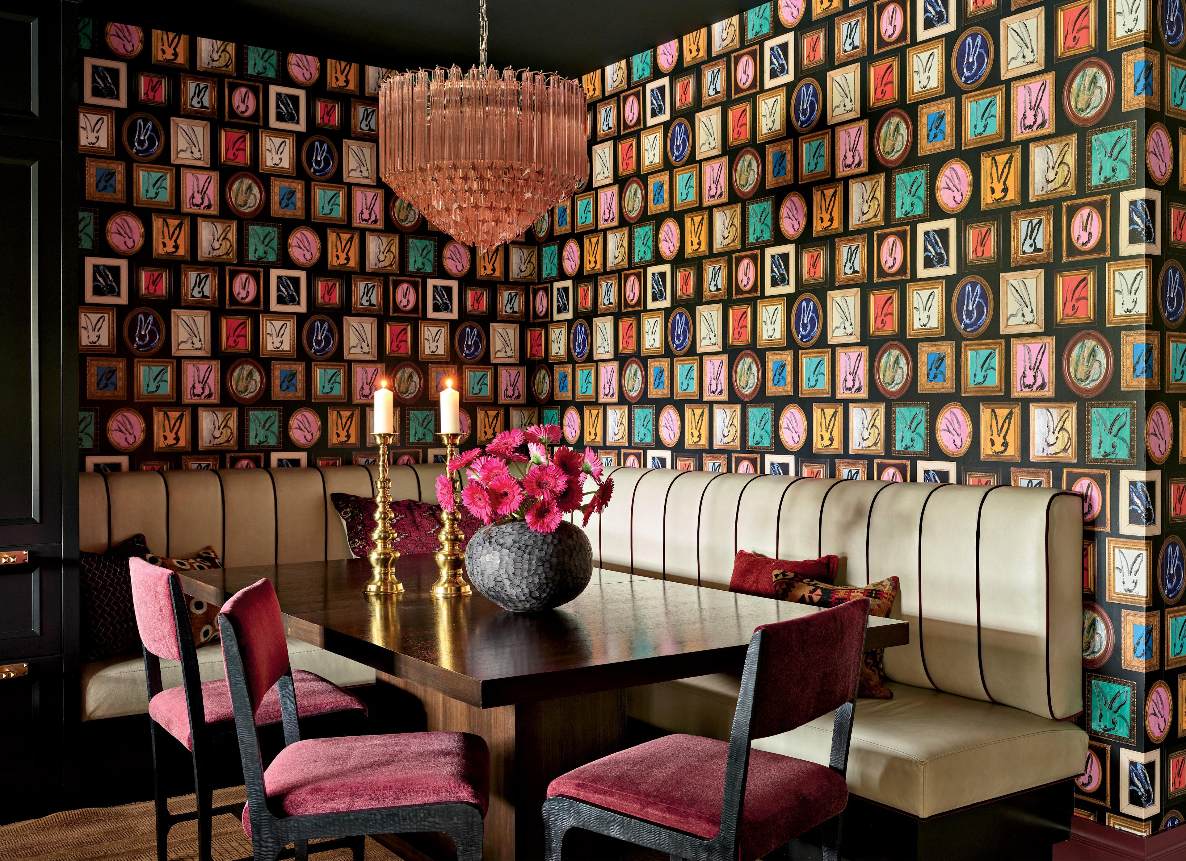 Dining room with colorful patterned wallpaper featuring framed images, a wooden table, pink-upholstered chairs, a plush beige bench, and an elegant chandelier.