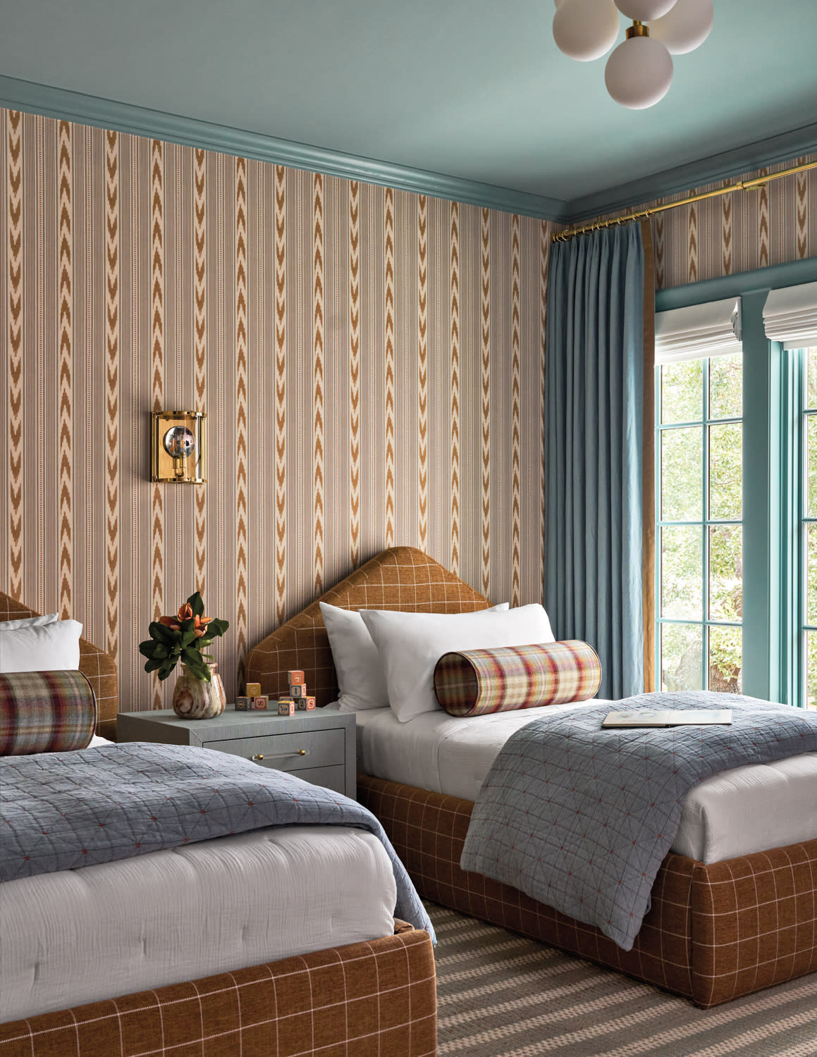 Two twin beds with plaid bolsters and blue quilts in a cozy bedroom. Patterned walls, floral centerpiece, and large window create a warm, inviting atmosphere.