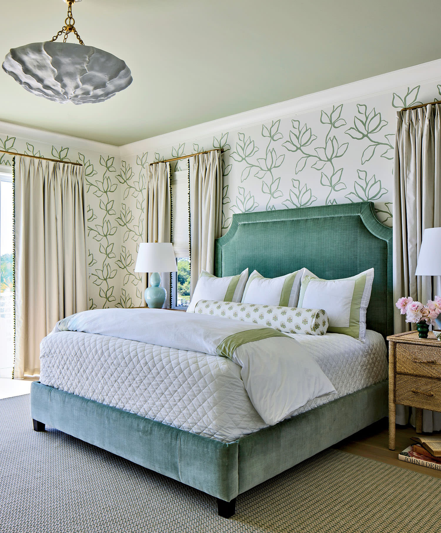 Elegant bedroom with a teal upholstered bed, white and green bedding, floral wallpaper, light curtains, textured ceiling fixture, and serene decor.