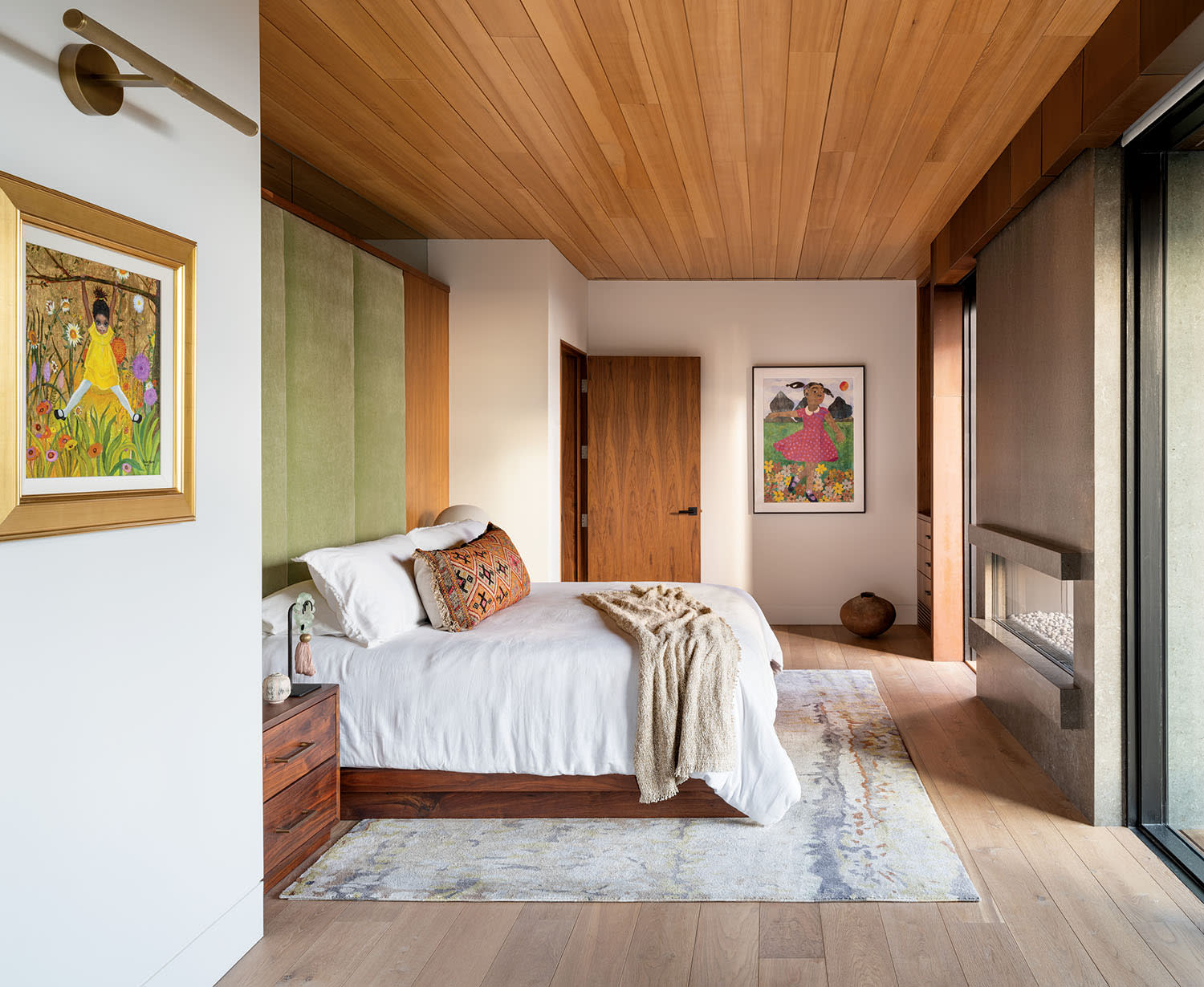 Modern bedroom with a wooden ceiling, white bed, and plush pillows. Vibrant artwork adorns the walls. Large window offers natural light and an inviting atmosphere.