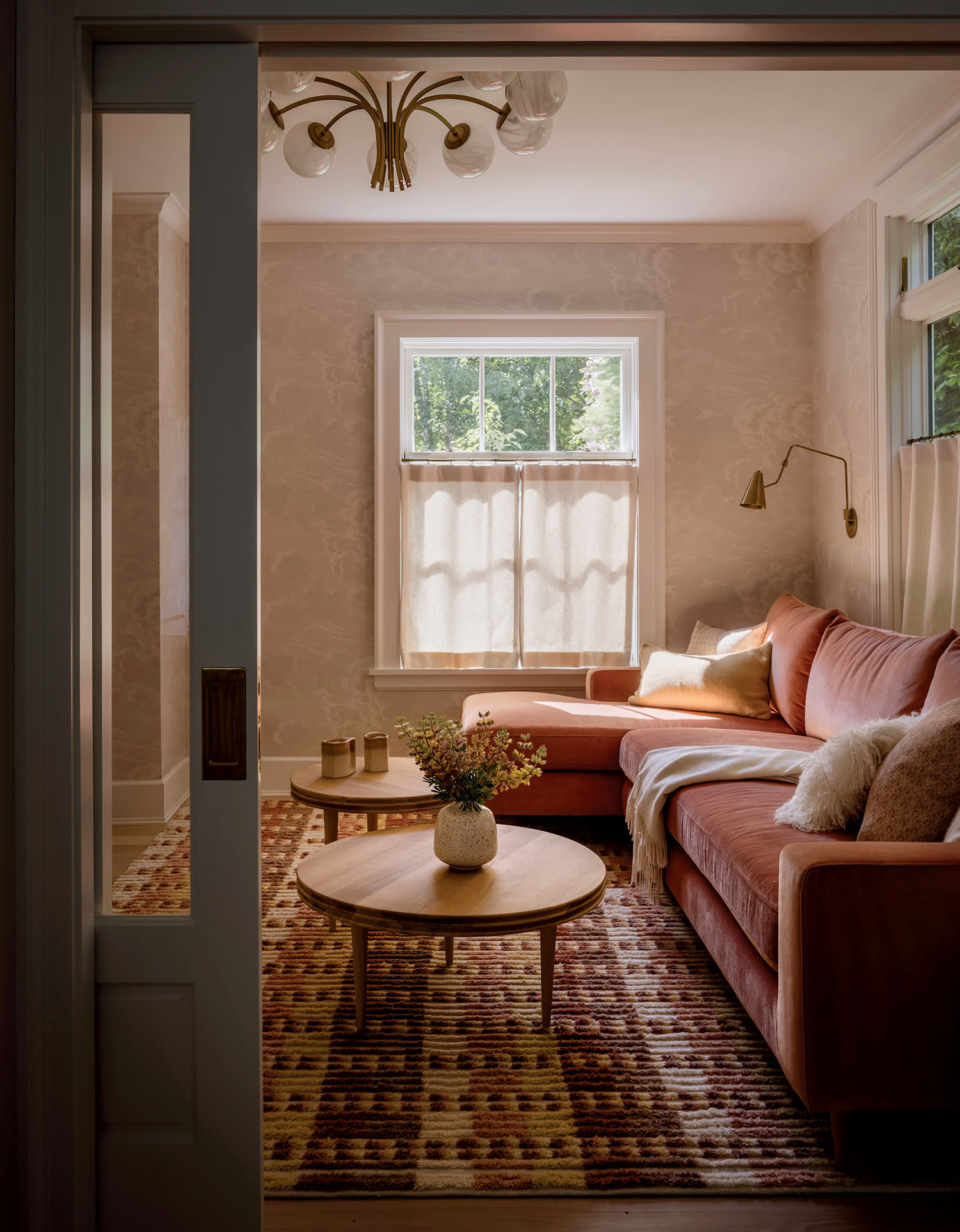 Cozy living room with a pink sofa adorned with pillows and a blanket. A round wooden table with a vase of flowers is on a patterned rug.