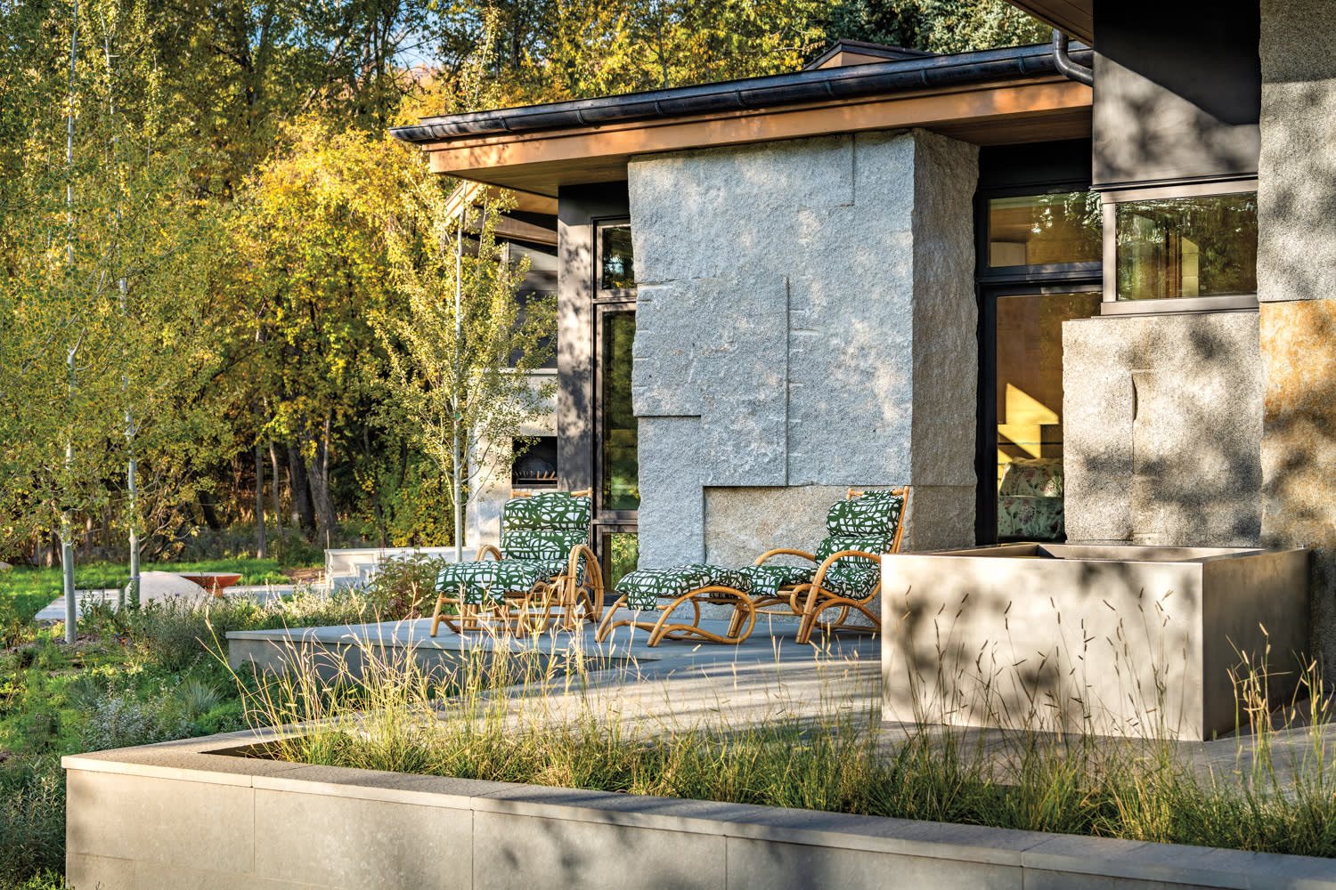 Exterior shot of the back of a house with a granite wall, natural-looking grasses and two rattan lounge chairs in a green-patterned fabric