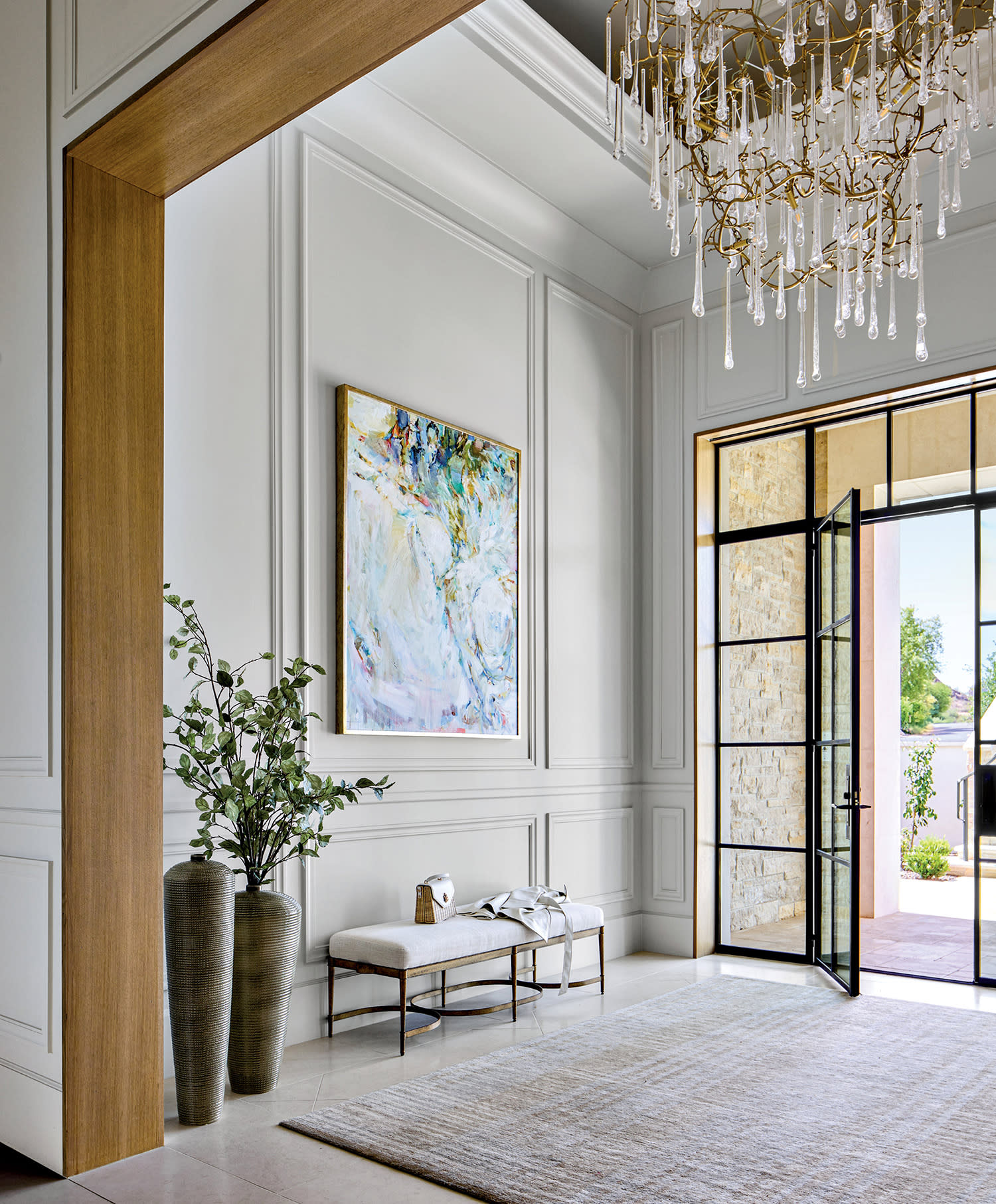 Luxurious foyer with a large crystal chandelier, abstract art, and potted plants. Open glass door reveals a stone wall outside, creating an elegant, airy feel.