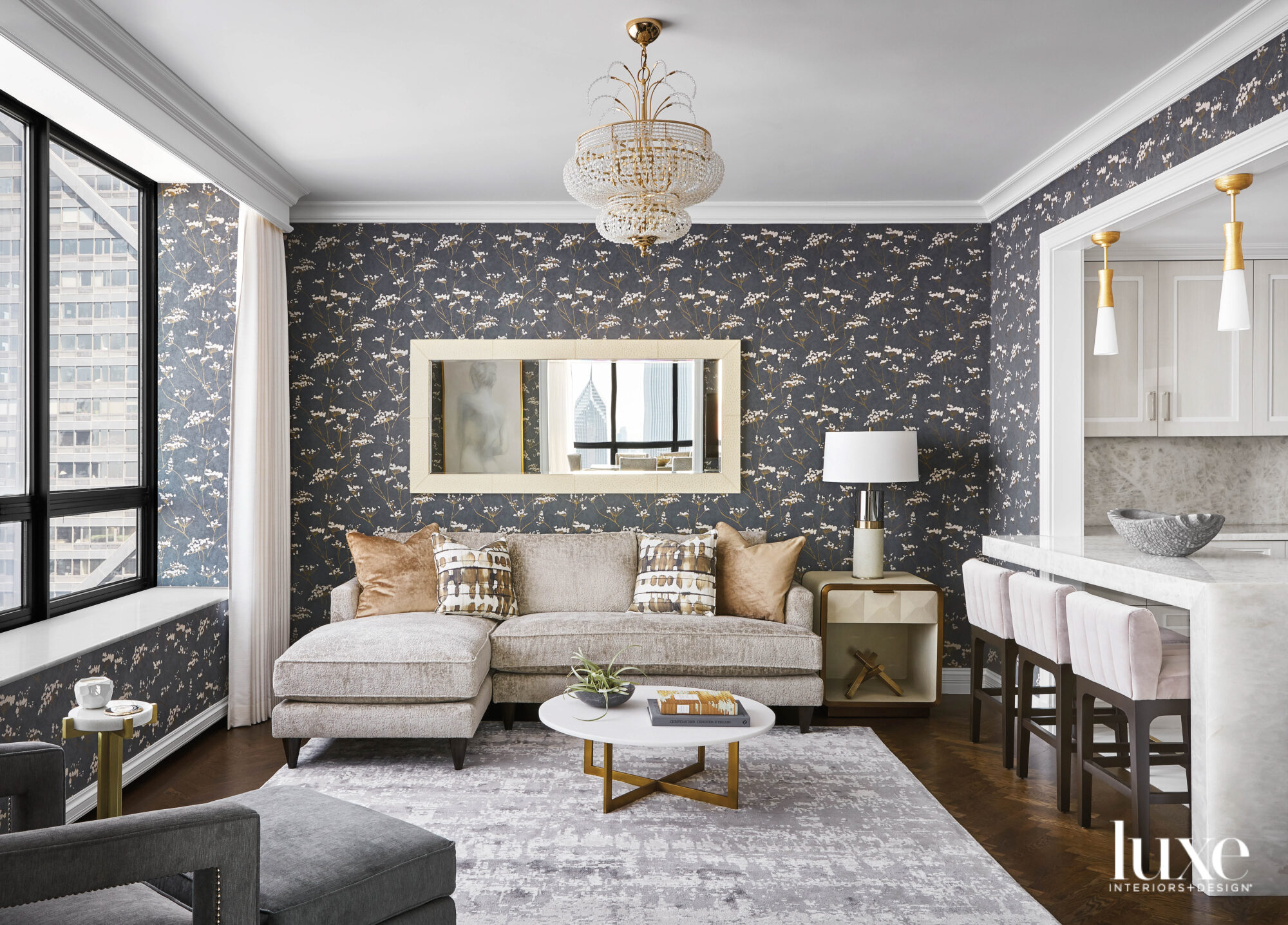 A tan sectional is in seating area off a kitchen. There is a dark floral wallpaper in the room.