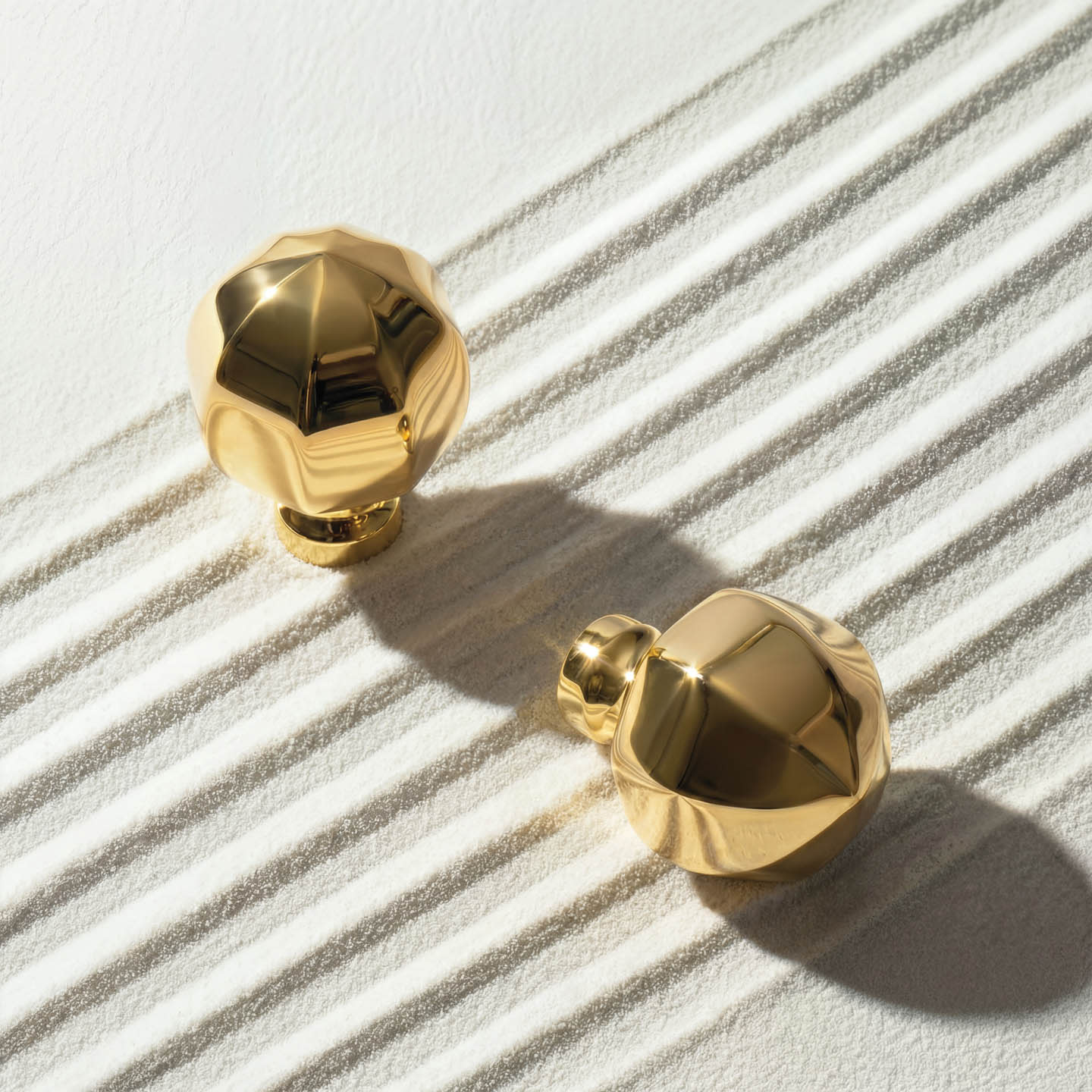 Two gold, faceted cabinet knobs on a textured white surface. Sunlight casts diagonal shadow lines, creating a luxurious and elegant atmosphere.