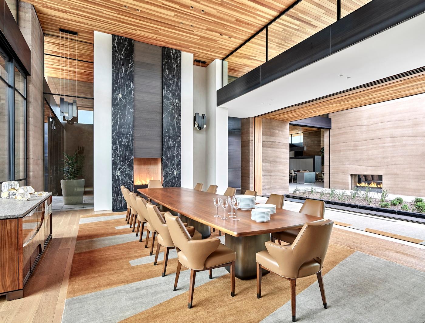 Modern dining room with a long wooden table, surrounded by beige chairs. A tall, black marble fireplace stands at one end. Glass doors open to a courtyard.