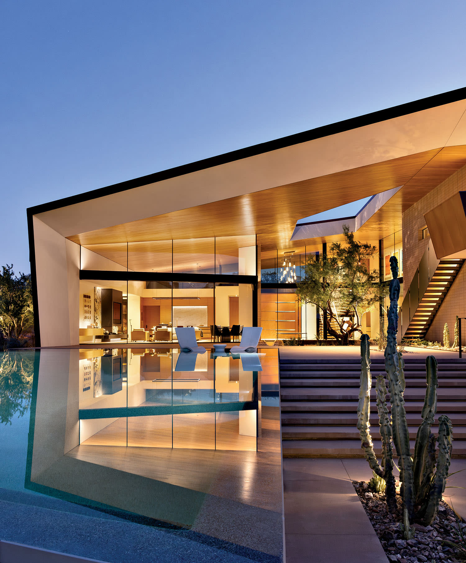 Modern glass-walled house at dusk with warm interior lighting, reflecting in an adjacent pool. Steps lead to the patio; a cactus stands nearby, enhancing the serene atmosphere.