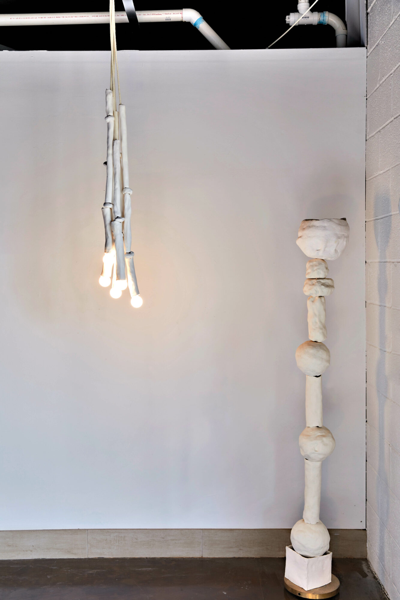 A white room with a tall standing sculpture and a dancing ceiling sculpture, illuminated by bulbs