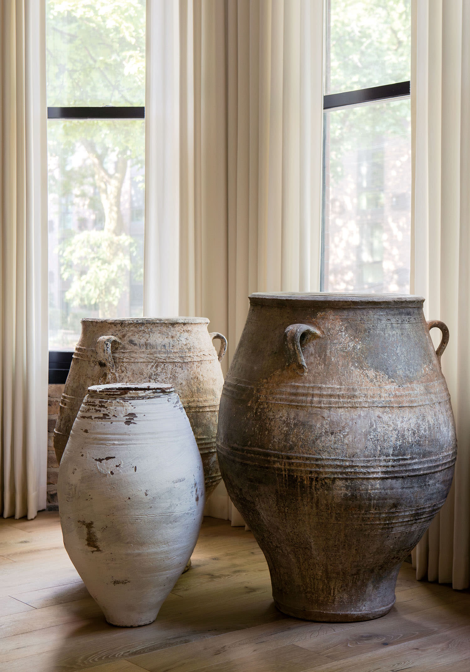 A set of three large vases arranged on the floor in a corner near two windows with opened curtains.