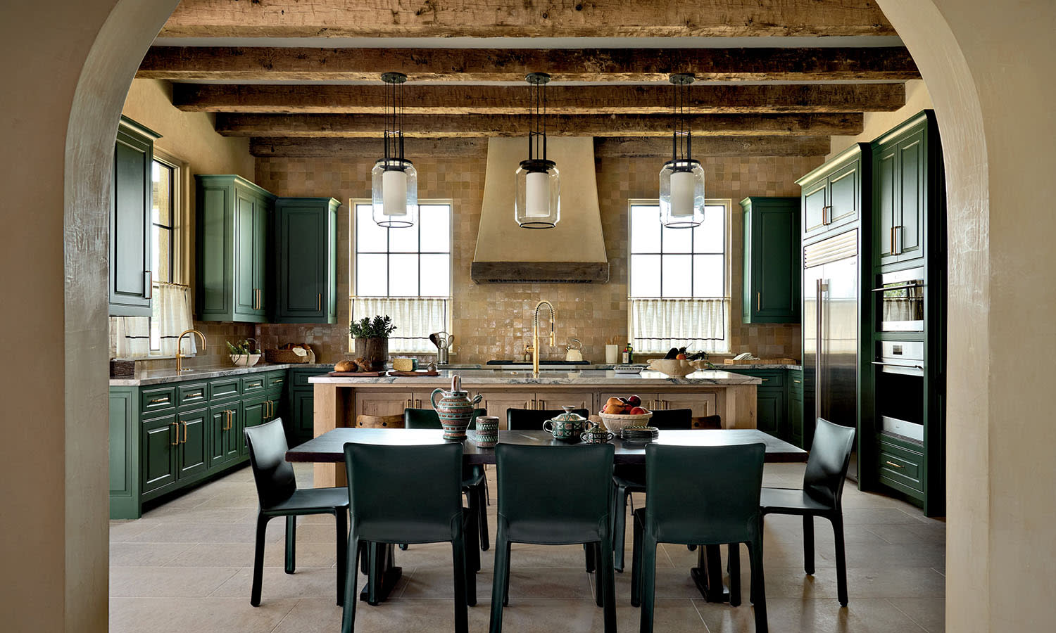 Rustic kitchen with green cabinets, exposed wooden beams, and hanging lights. A large table with six chairs centers the space, exuding warmth and elegance.