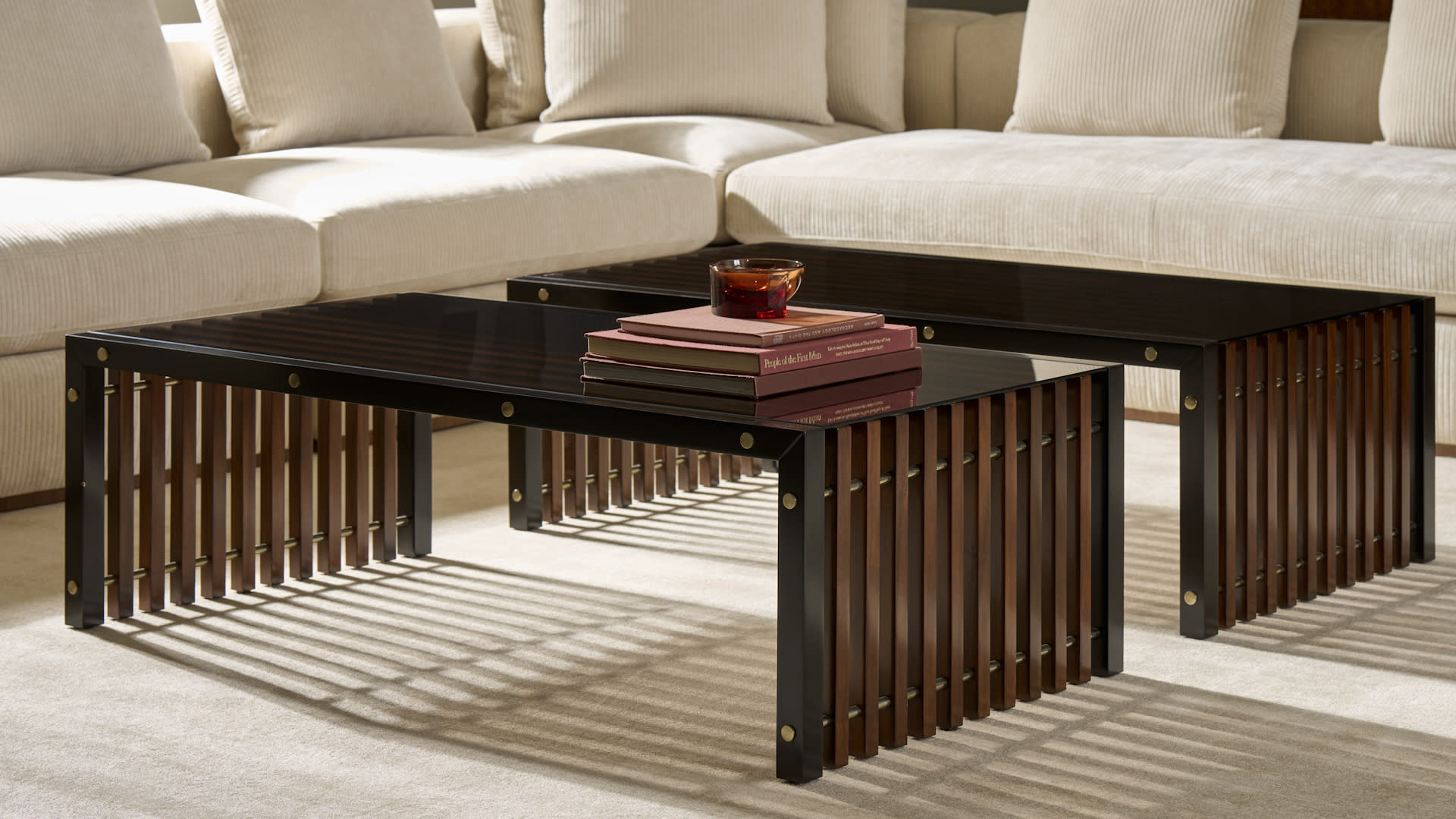 Two modern coffee tables with black tops and wooden slatted sides on a beige carpet. Tables hold stacked books and a red bowl, near a cream sofa. Cozy ambiance.