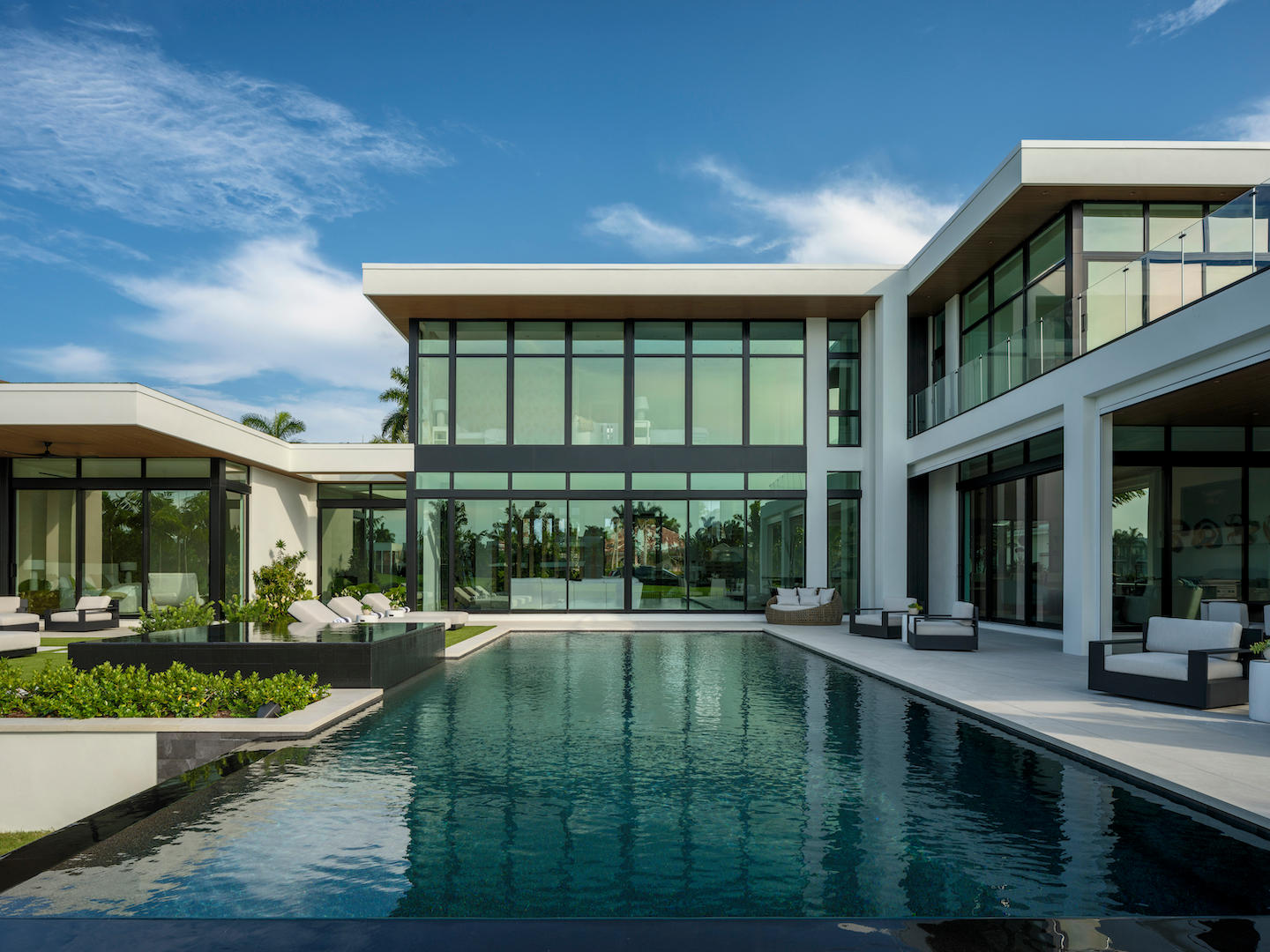 Contemporary waterfront estate clad in white stucco and expansive black-framed glass, with double-height windows and floating rooflines reflecting a vivid blue sky. A sleek, resort-style lap pool anchors the courtyard, flanked by cushioned loungers, modern patio seating, and low planters of lush tropical greenery.
