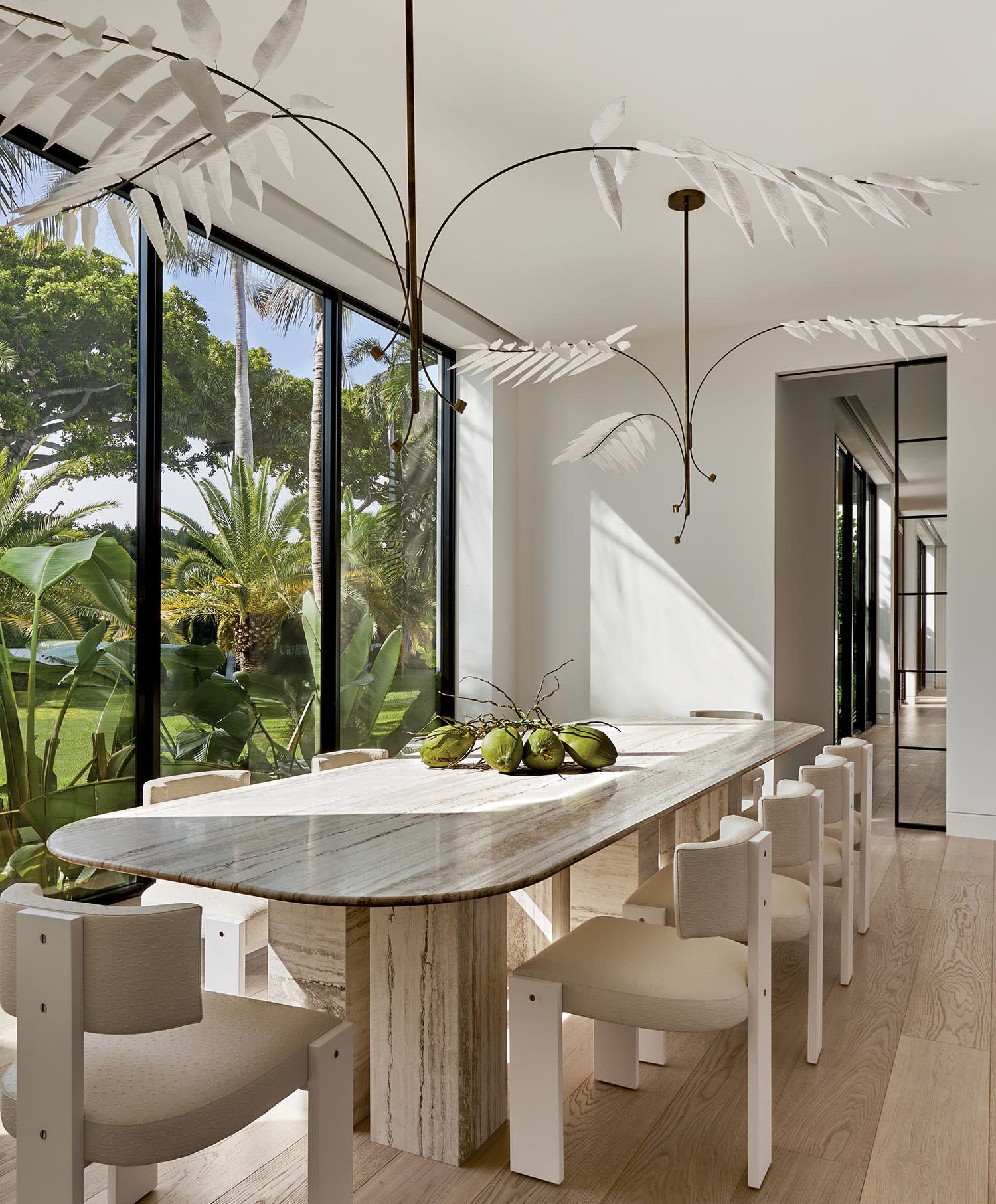 Stylish dining room with a long marble table, surrounded by white chairs. A modern chandelier hangs above. Large windows reveal lush greenery outside.