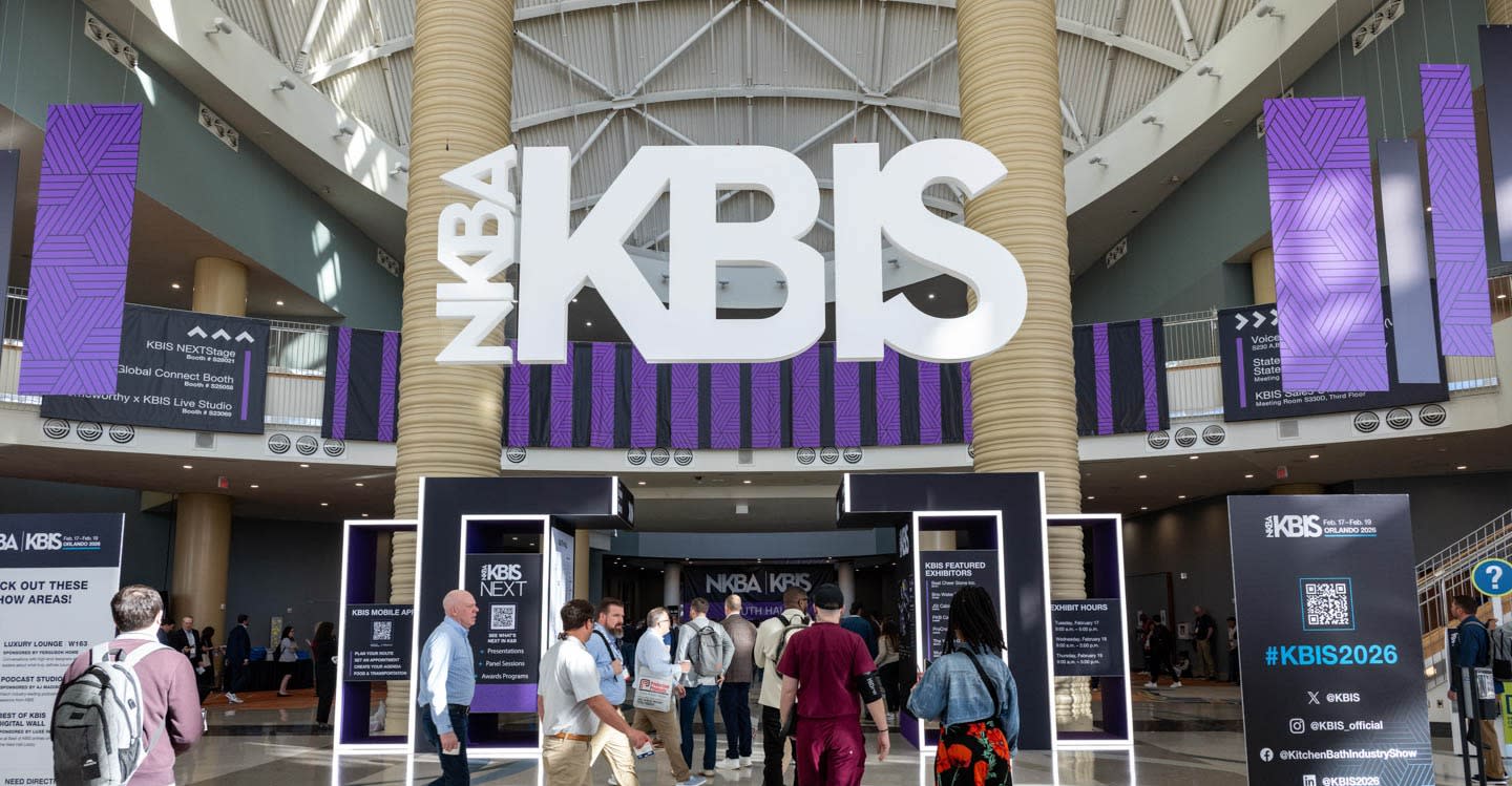 People walk under a large "KBIS" sign at a convention center entrance. The atmosphere is lively, with purple banners and event displays in the background.