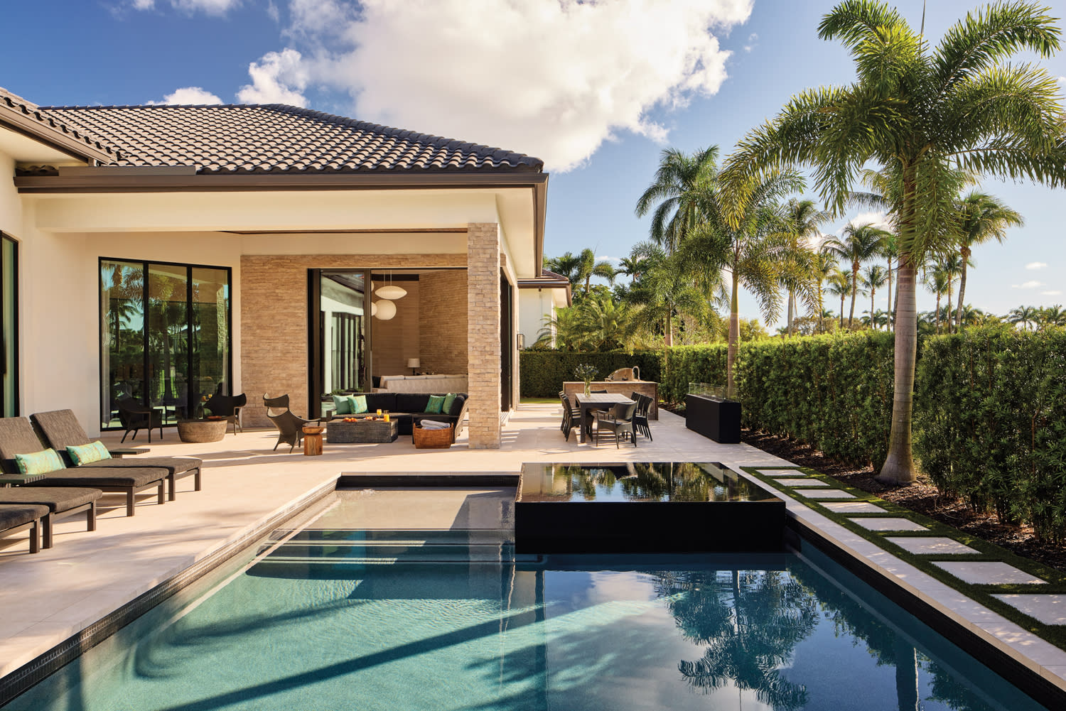 Modern backyard showing a luxurious patio with lounge chairs, seating area, dining table, and pool. Palm trees and hedges surround the sunny scene.