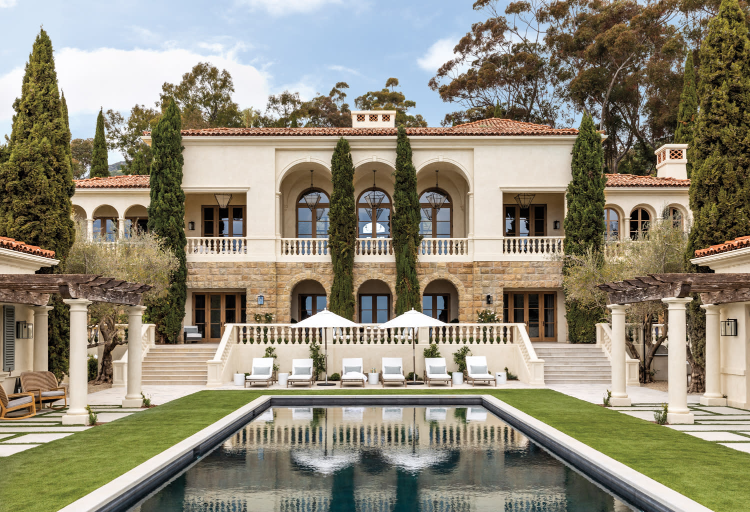 Back facade of mediterranean style home, with large rectangular pool set and white chaise loungers backed by Italian cypress trees