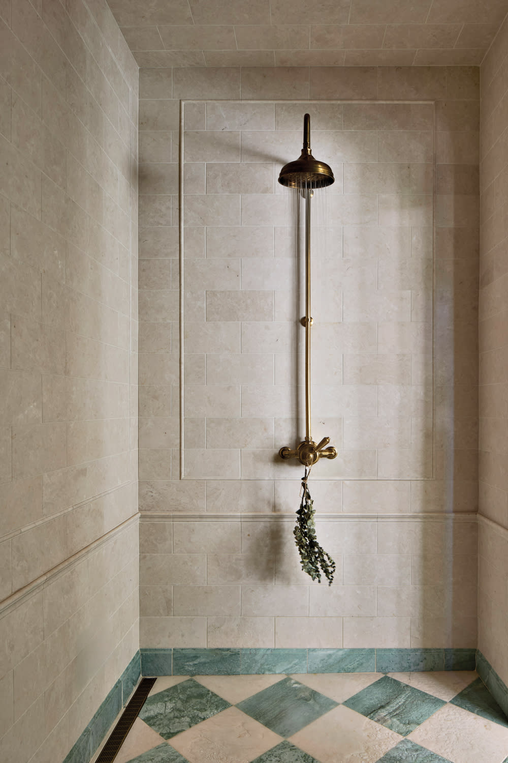 Elegant shower with a vintage bronze fixture on beige stone tiles. Green and beige checkered floor; tied herbs hang from the shower handle, creating a serene, spa-like ambiance.