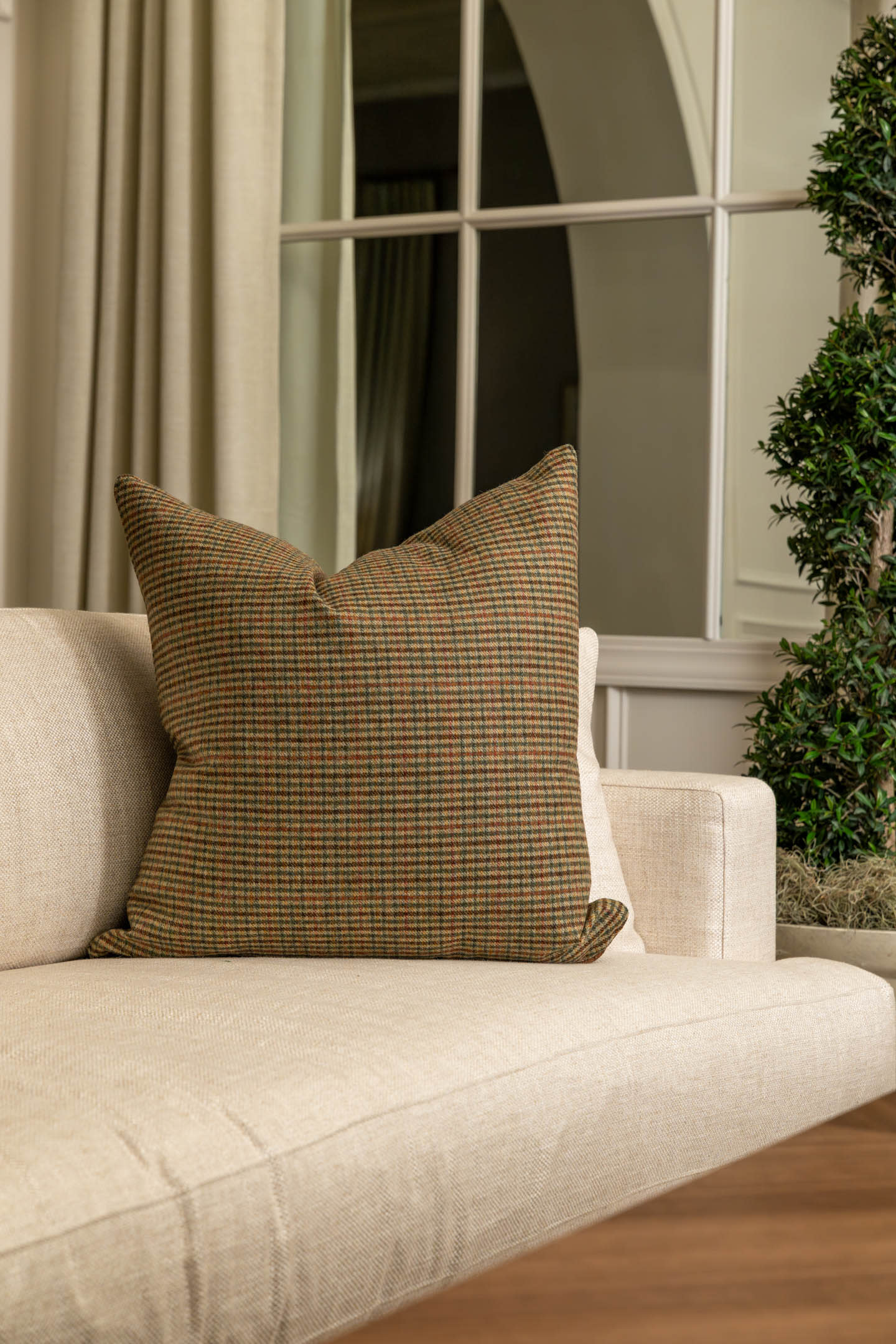 A cozy, light beige sofa features a houndstooth-patterned cushion in green and brown tones. Behind are floor-length curtains and a large window, adding elegance.