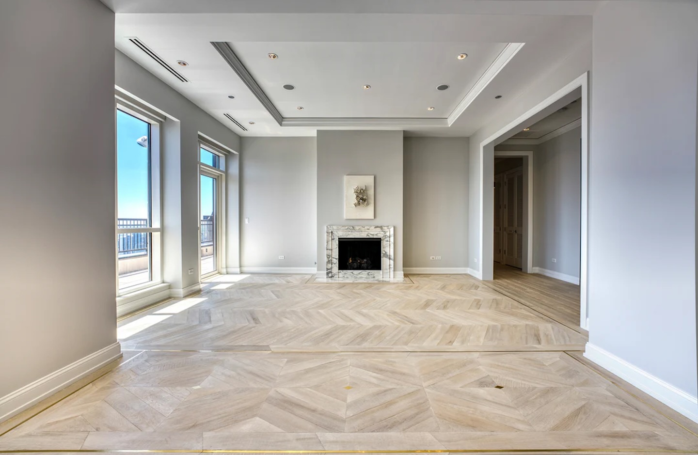 Spacious and elegant living room with light wood flooring, a marble-framed fireplace, and large windows offering vibrant natural light and city views.