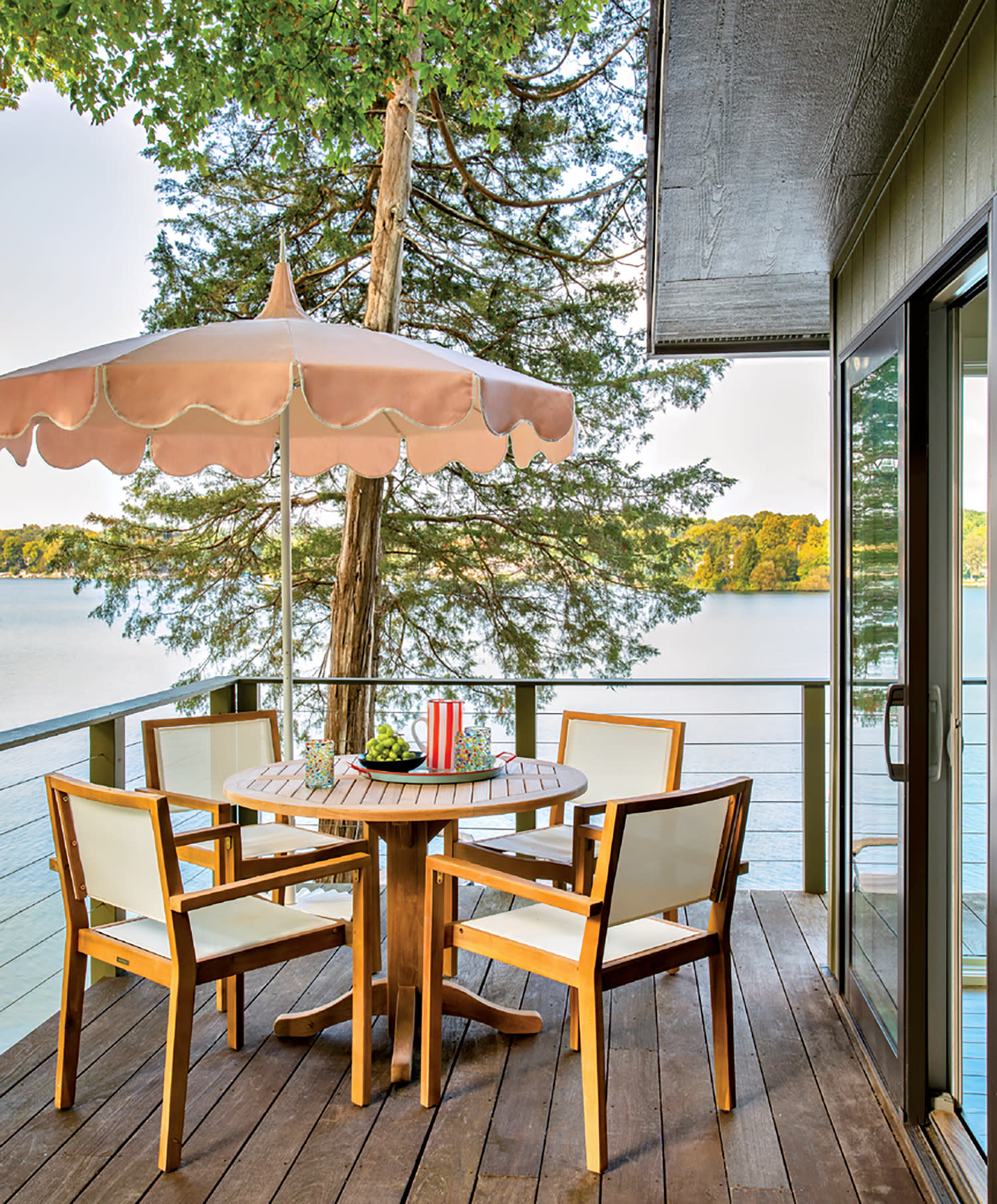 A serene lakeside deck features a wooden table with four chairs under a pink umbrella. A tree provides shade, while the calm water creates a tranquil setting.