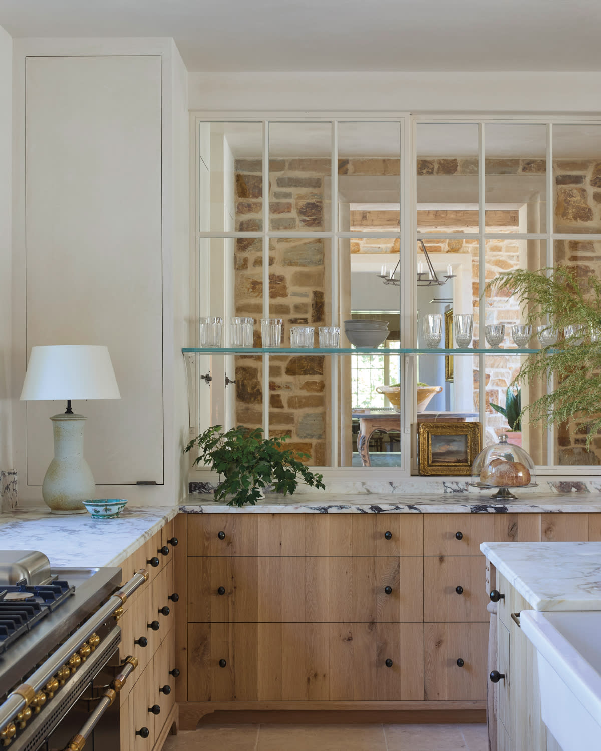 Cozy kitchen with light wood cabinets and marble countertops. A large window with a shelf holds glassware, framing a view of a rustic dining area.