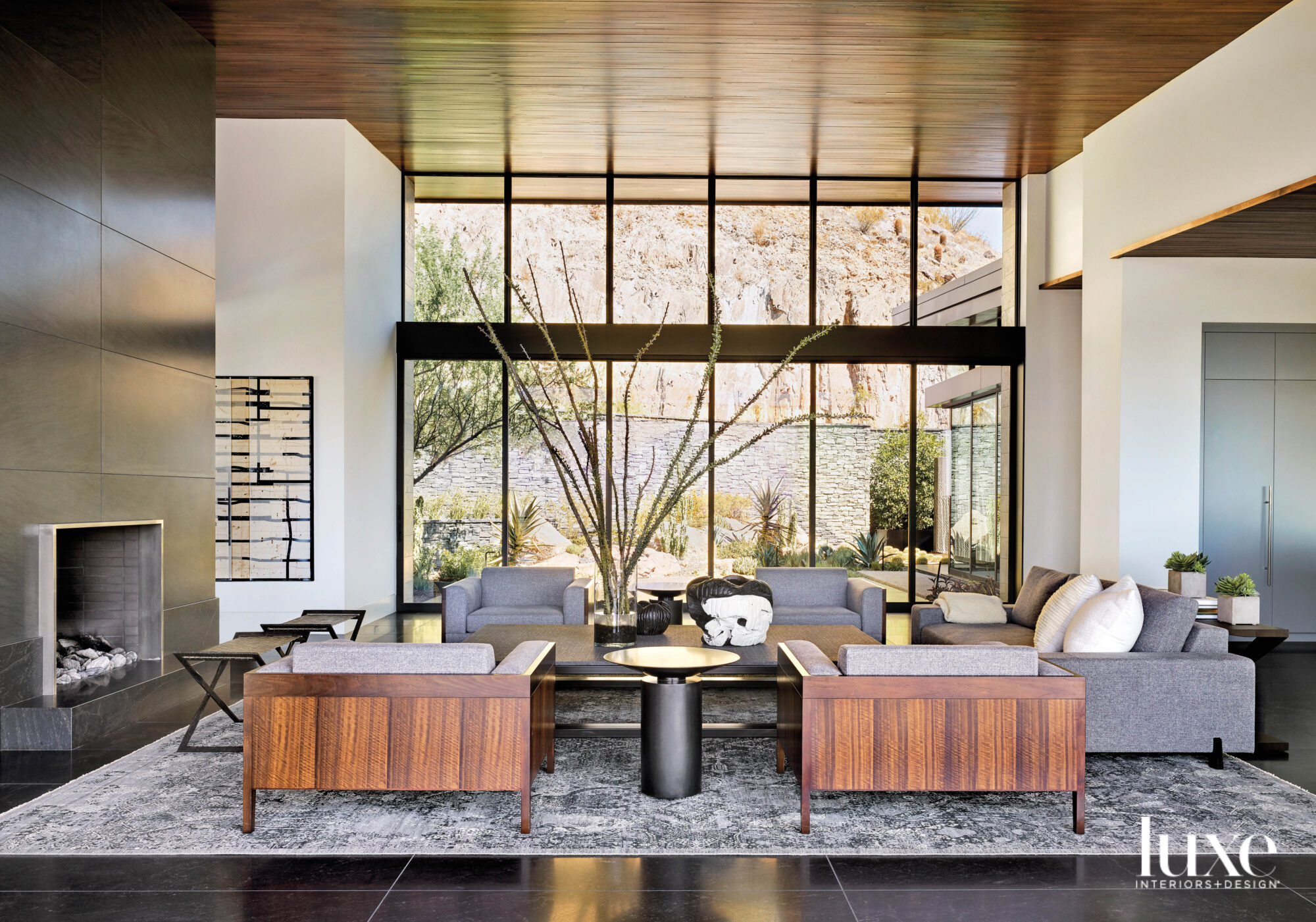 A living room with a floor-to-ceiling stone fireplace, gray and wood furnishings, and a granite floor. Floor-to-ceiling windows look out on a desert garden.