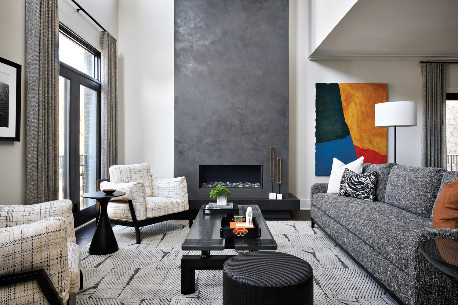 A living room with a gray plaster fireplace, a gray couch, two white plaid chairs and a yellow, red, blue and black painting next to the fireplace.