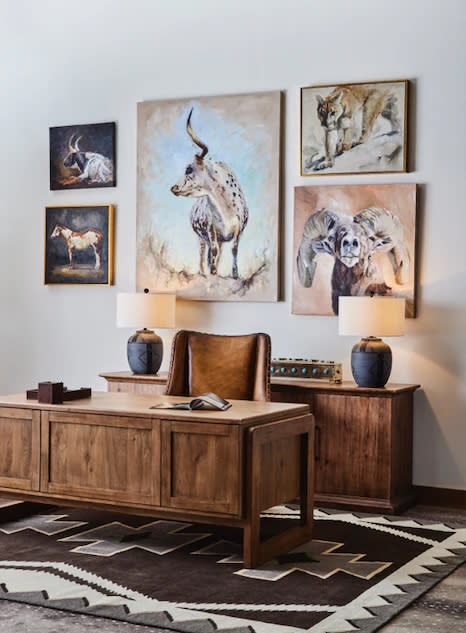 A rustic office features a wooden desk with a leather chair, flanked by two table lamps. Animal artwork, including cows and an elephant, adorns the wall, set on a geometric-patterned rug, conveying a warm, natural ambiance.