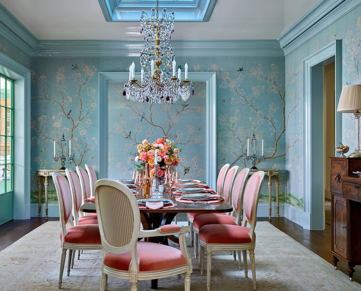 Elegant dining room with a pastel blue theme, featuring a chandelier, floral wallpaper, pink cushioned chairs, and a skylight. A centerpiece of colorful flowers on the table adds vibrancy.
