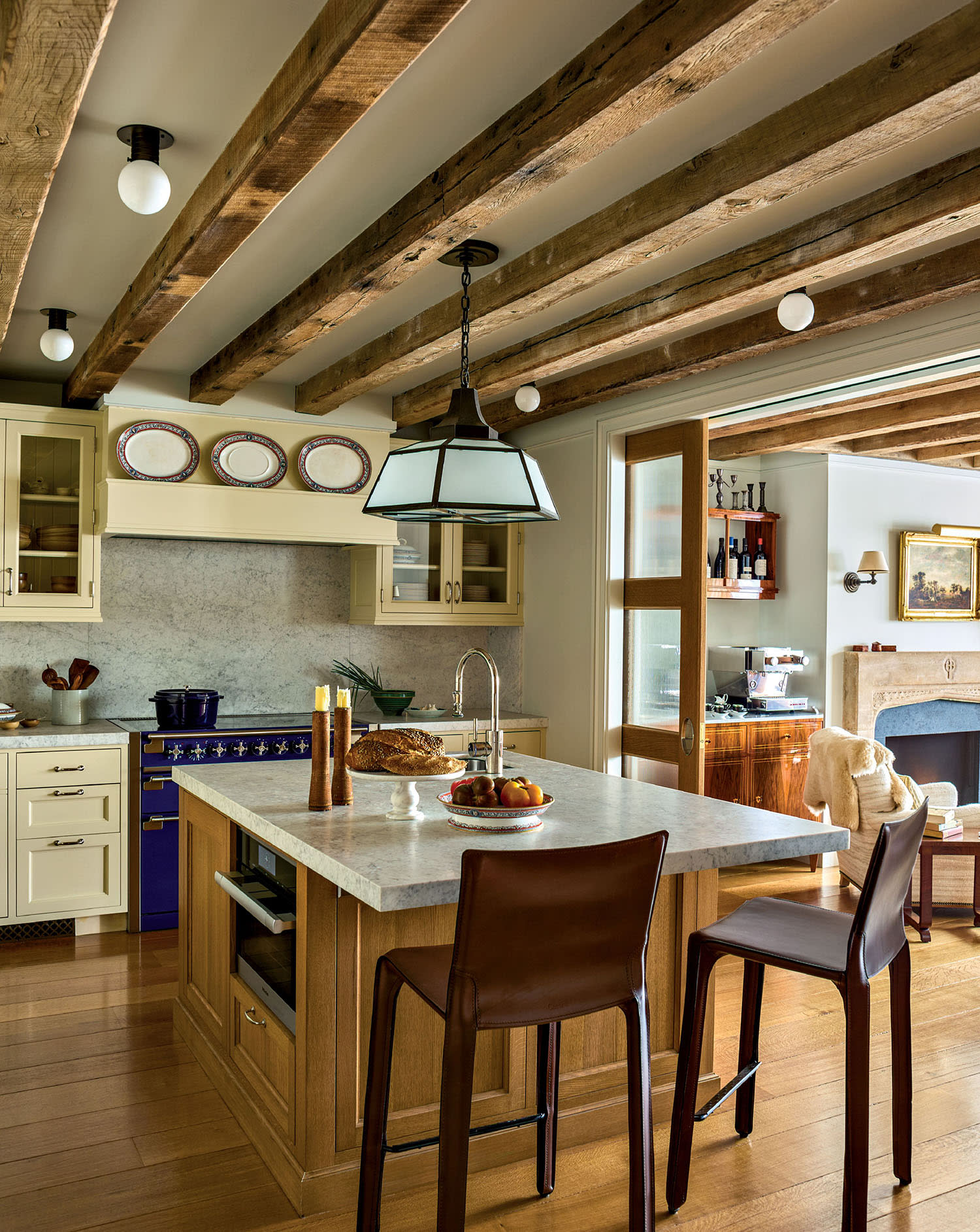Stylish kitchen with wooden beams, marble island, and dark blue stove. Brown chairs add warmth, while pendant lamp creates a cozy, inviting ambiance.
