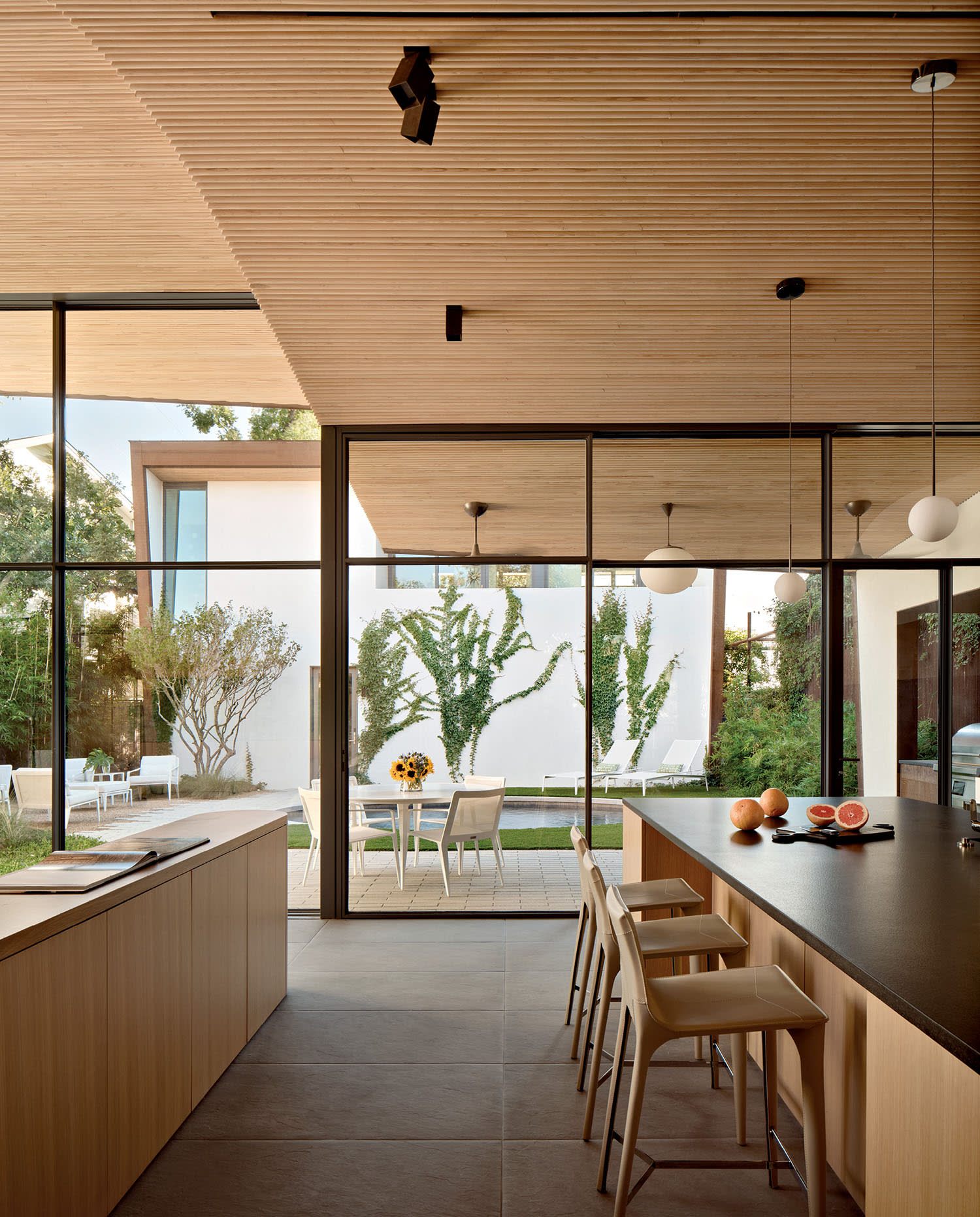 Modern kitchen with wood accents features a large island and bar stools. Glass doors open to a patio with greenery, creating a serene atmosphere.