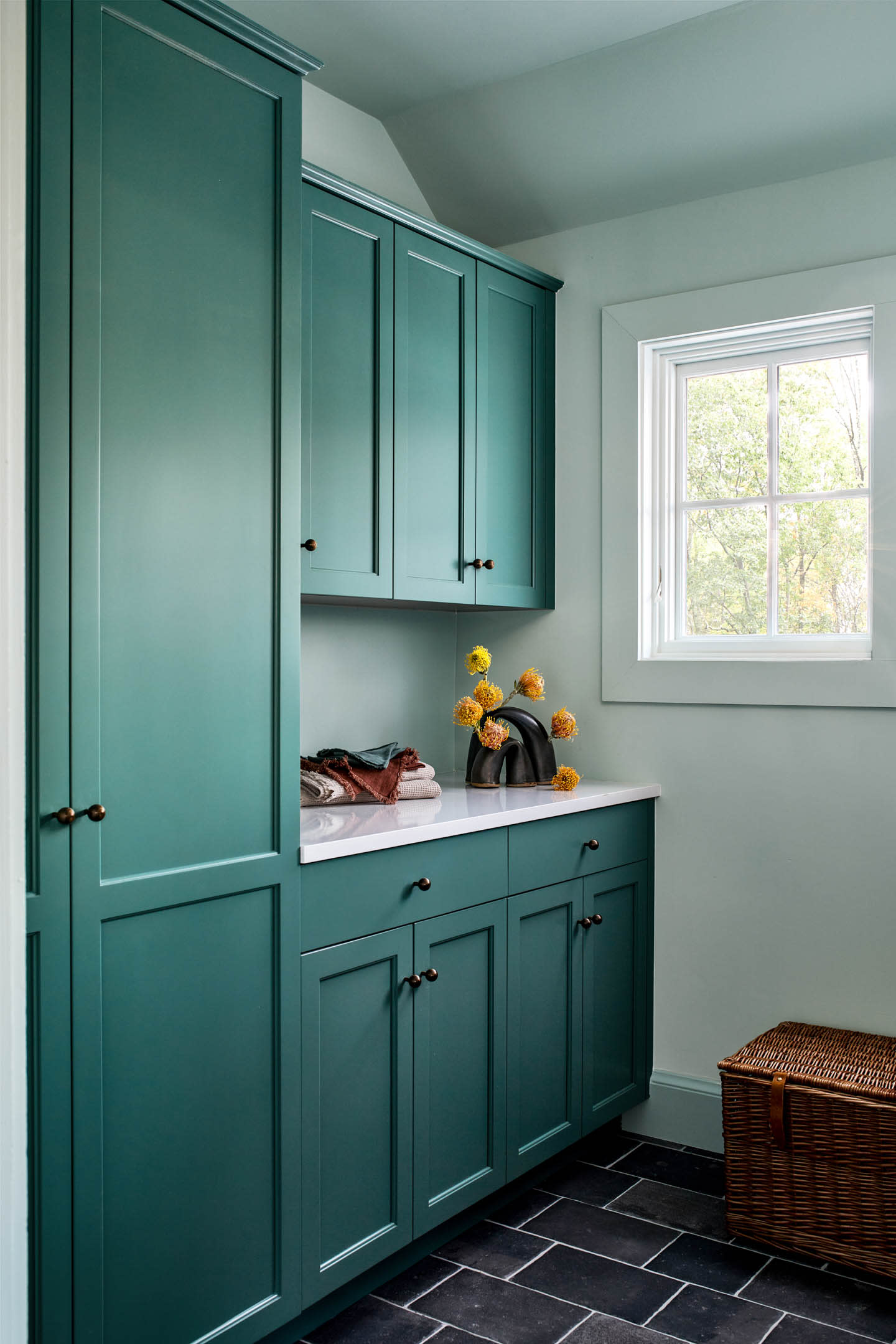 A bright laundry room with teal cabinets, a white countertop, and a vase of yellow flowers. Sunlight filters through a window, creating a fresh, inviting atmosphere.