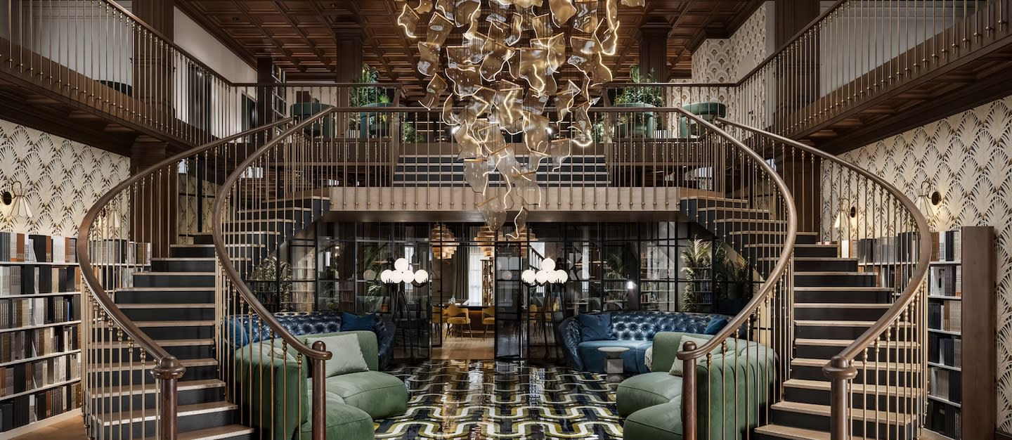 Elegant living room with a grand double staircase, modern chandelier, and luxurious seating. Sophisticated, warm ambiance with geometric patterned floor.