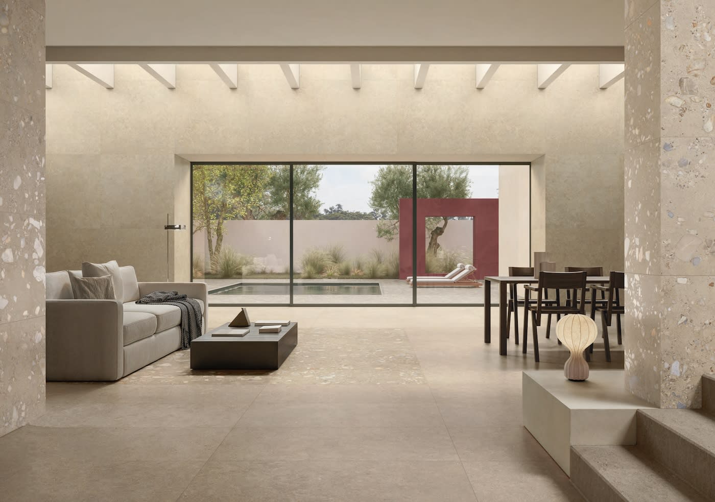 Spacious modern living room with beige stone walls, a gray sofa, and a minimalistic dining set. Large windows overlook a serene garden and pool.