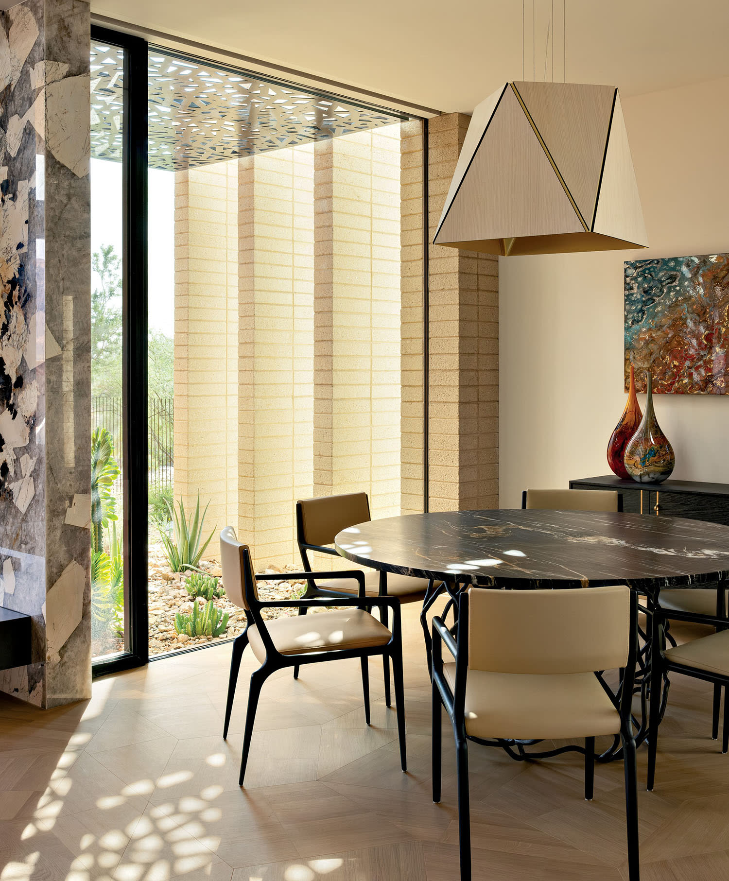 Modern dining room with a round black stone table, beige chairs, and geometric pendant light. Large windows illuminate colorful art and vases.