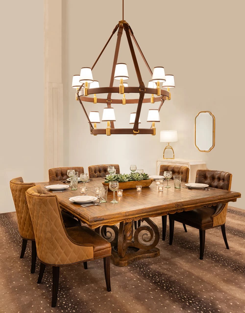 Elegant dining room with a wooden table set for six, surrounded by brown tufted chairs. A large chandelier hangs above, casting warm light. A console table with a lamp and mirror is in the background.