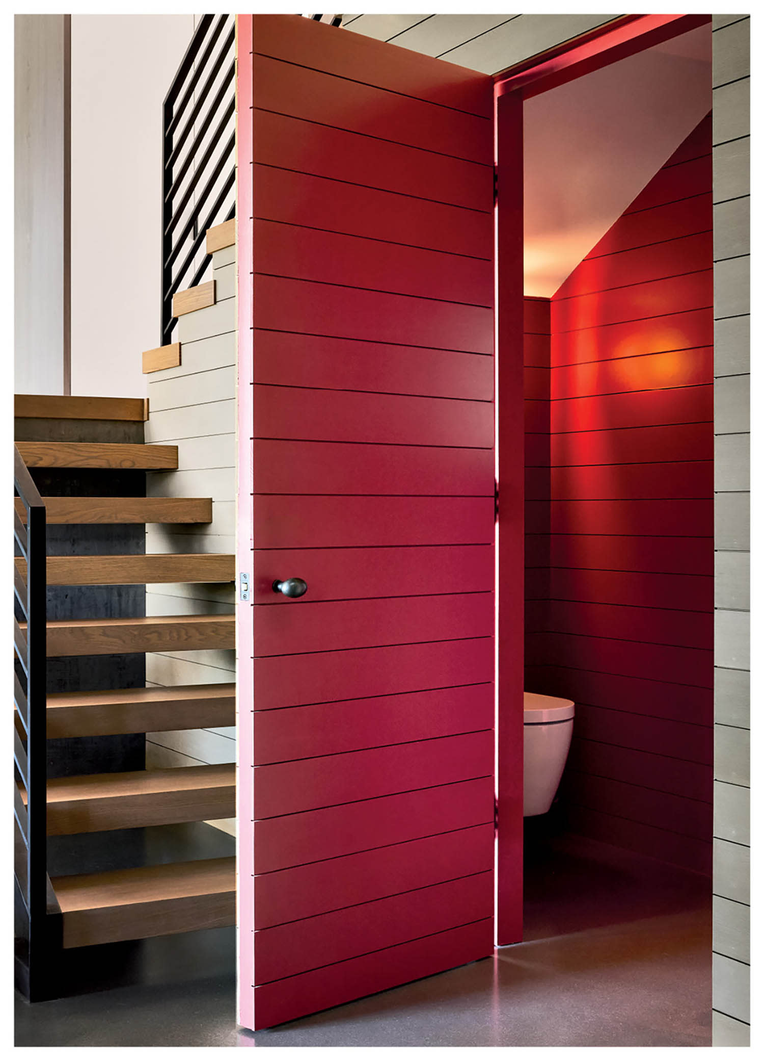 Staircase with wooden steps and black railing beside an open red door revealing a small bathroom. The scene feels modern and minimalistic.