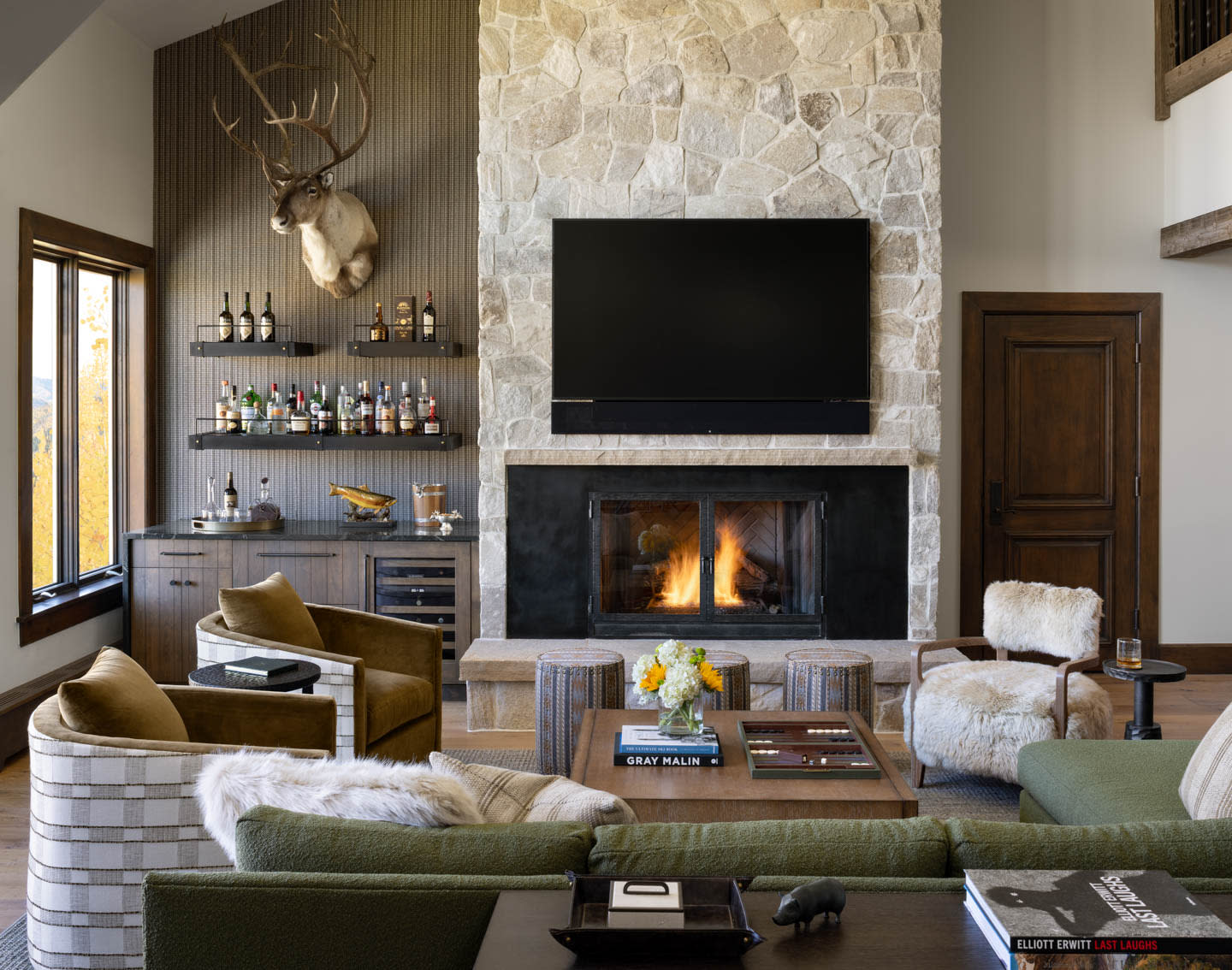 Cozy living room with a stone fireplace, mounted TV, and warm fire. Elegant decor includes a plush sofa, armchairs, and a wall-mounted deer head.