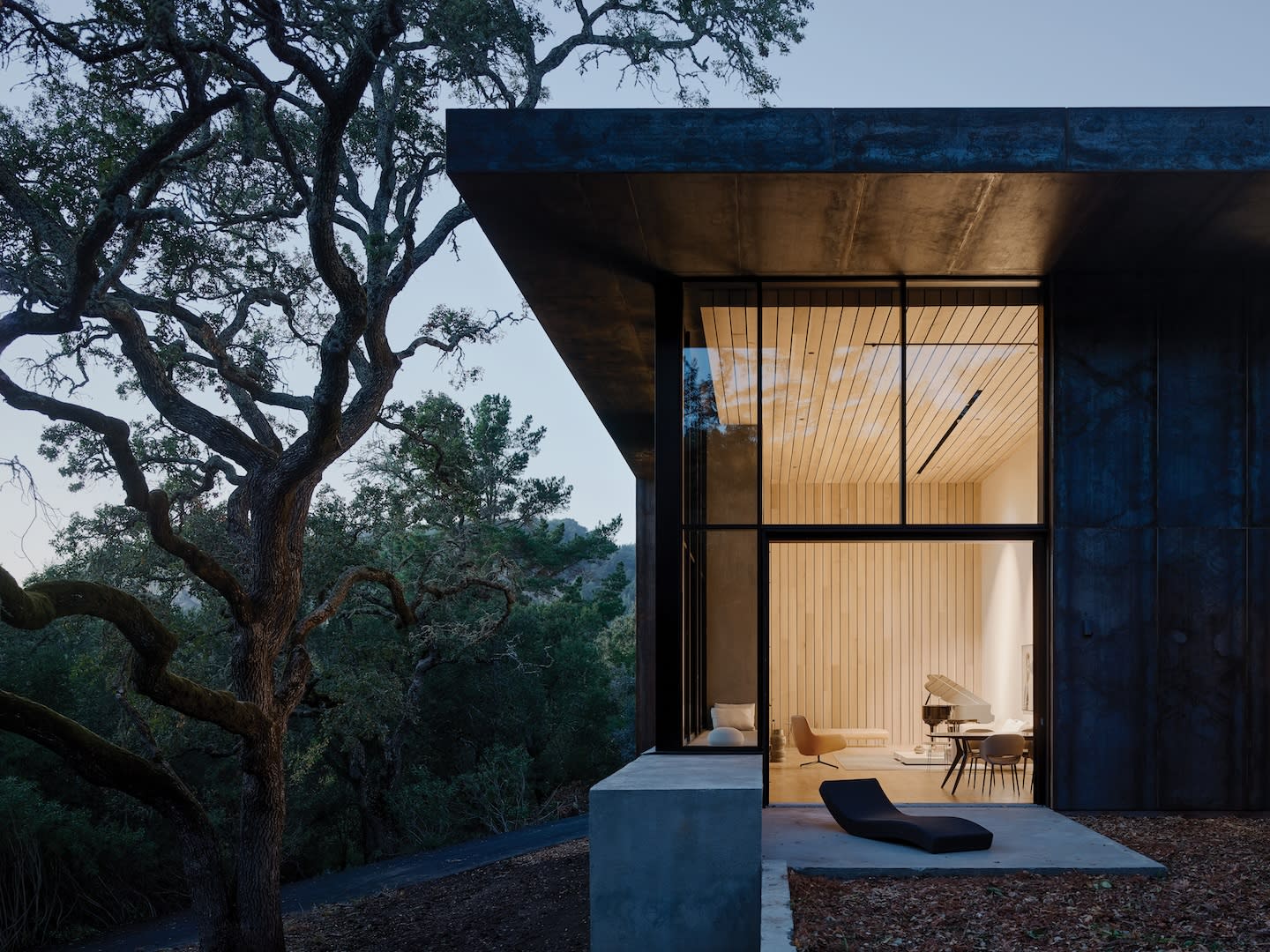 Modern home with large glass windows, revealing a warmly lit interior at dusk. Surrounded by trees, it conveys tranquility and seamless nature integration.