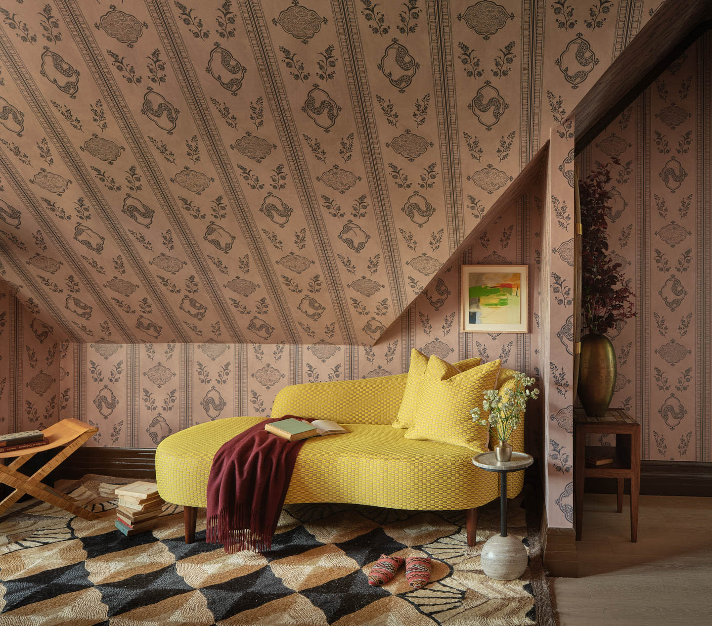 Cozy attic room with patterned wallpaper, featuring a yellow chaise with pillows, a small side table, books, and a colorful geometric rug.