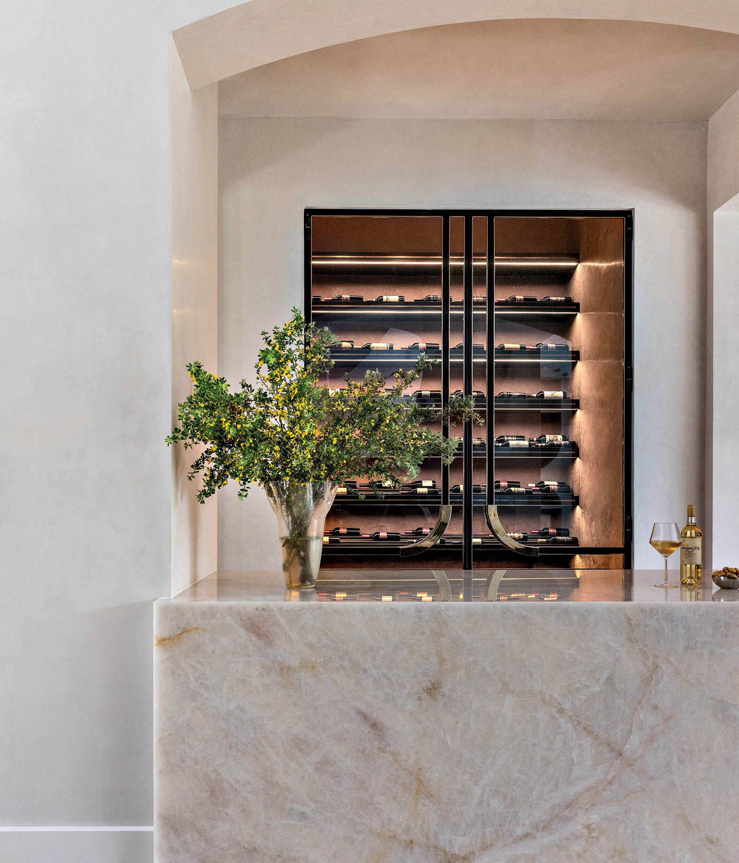 A stone wine bar sits in a home's entryway in front of a wine cellar with steel-and-brass doors.