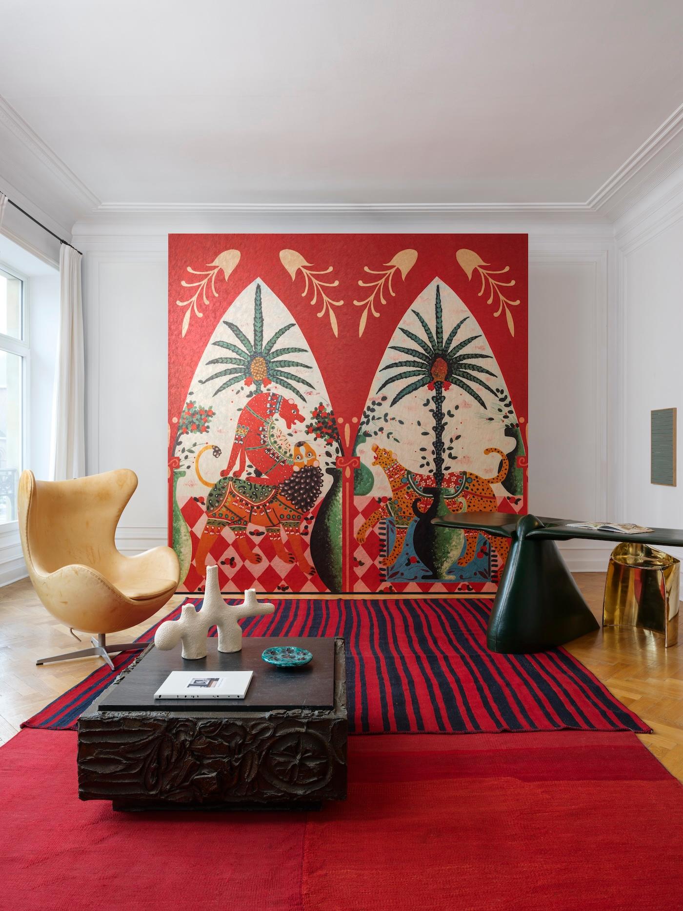 Modern living room with a striking red wall featuring colorful, artistic designs of figures and animals under palm trees. A yellow chair, stylish black table, and red-striped rug enhance the eclectic look. Bright, inviting atmosphere.