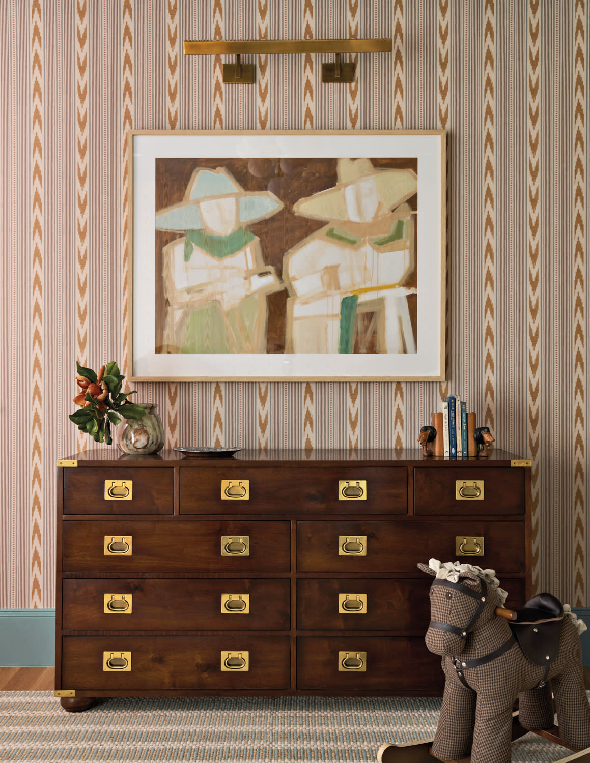 A brown wooden dresser with brass handles is set against a patterned wall. Above, a framed abstract painting hangs. A plush horse toy sits beside it.