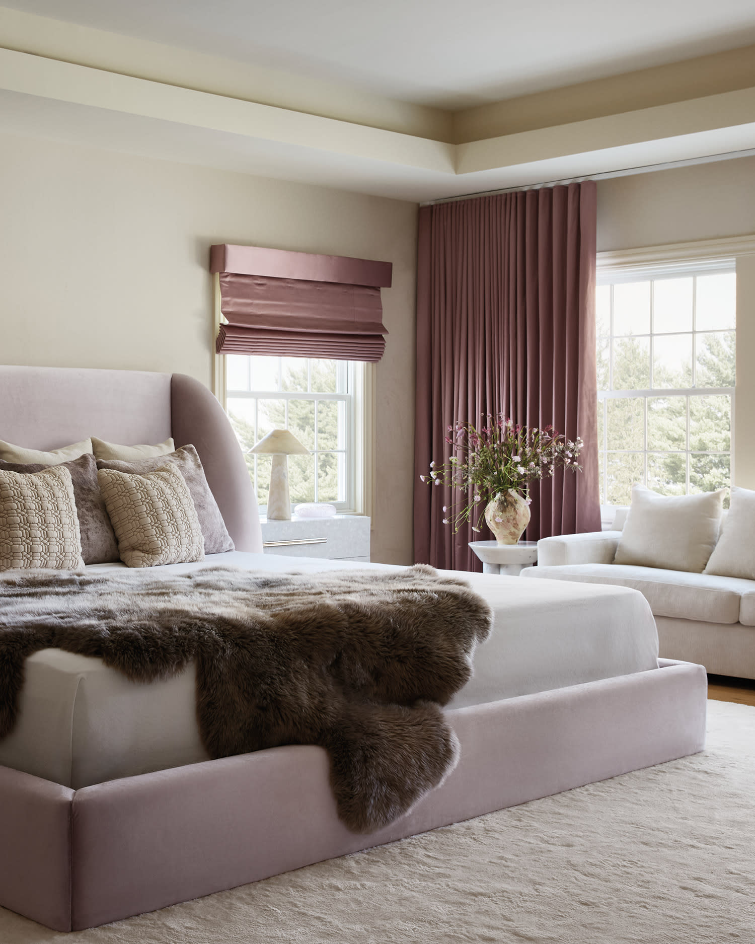 Lavender upholstered bed in a serene bedroom with pink drapes and cream seating.