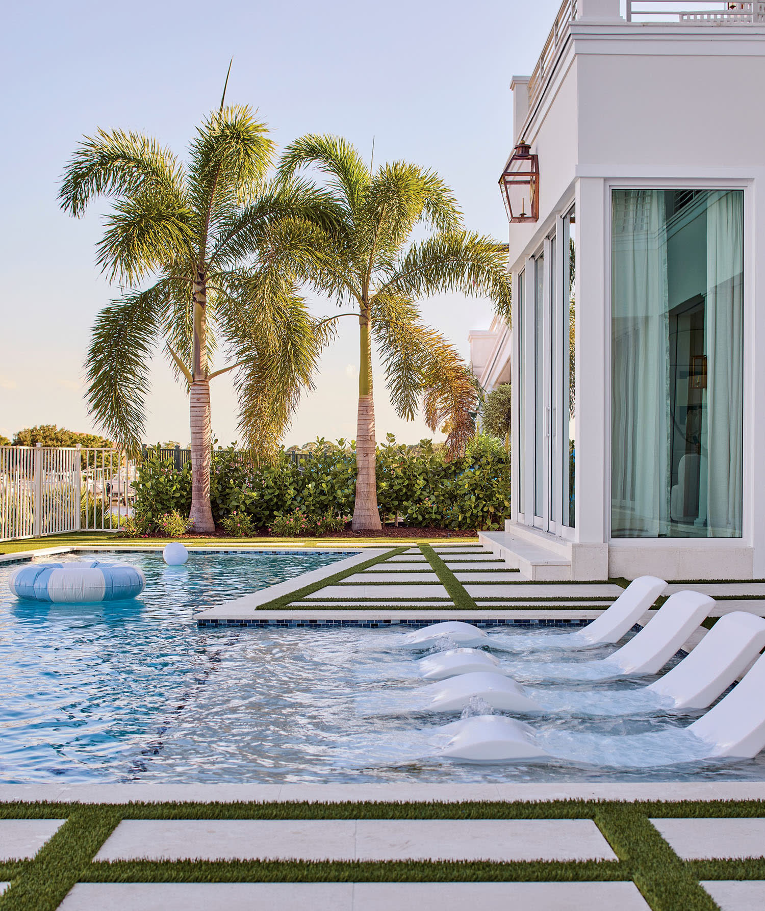 Luxurious poolside scene with three submerged loungers, tall palm trees, and a modern white building. The mood is serene and inviting under a clear sky.